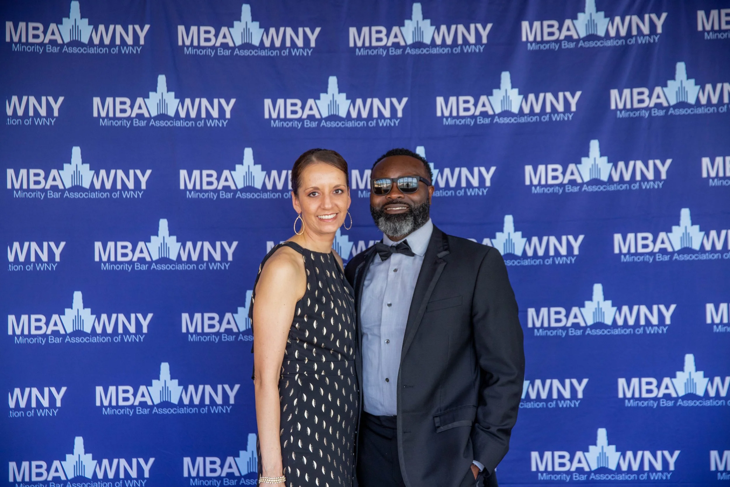 Two people standing in front of a blue backdrop with the text 'MBA WNY Minority Bar Association of WNY' repeated in a pattern. The woman on the left is wearing a sleeveless black dress with gold polka dots, gold hoop earrings, and a bracelet. The man