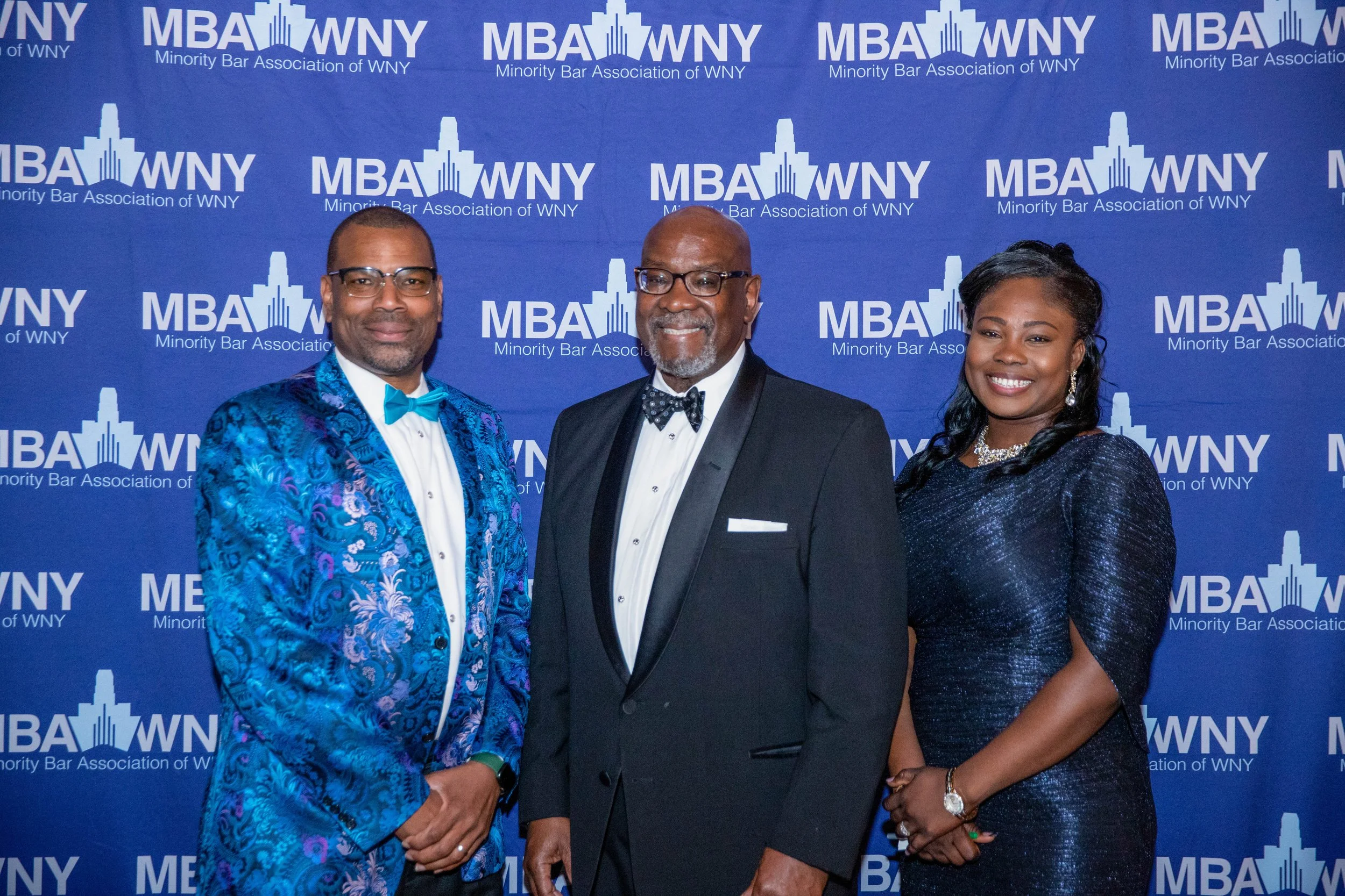 Three well-dressed individuals standing in front of a blue backdrop with white logos for the Minority Bar Association of WNY and MBA WNY. The person on the left is wearing a bright blue patterned suit with a matching bow tie. The person in the middle