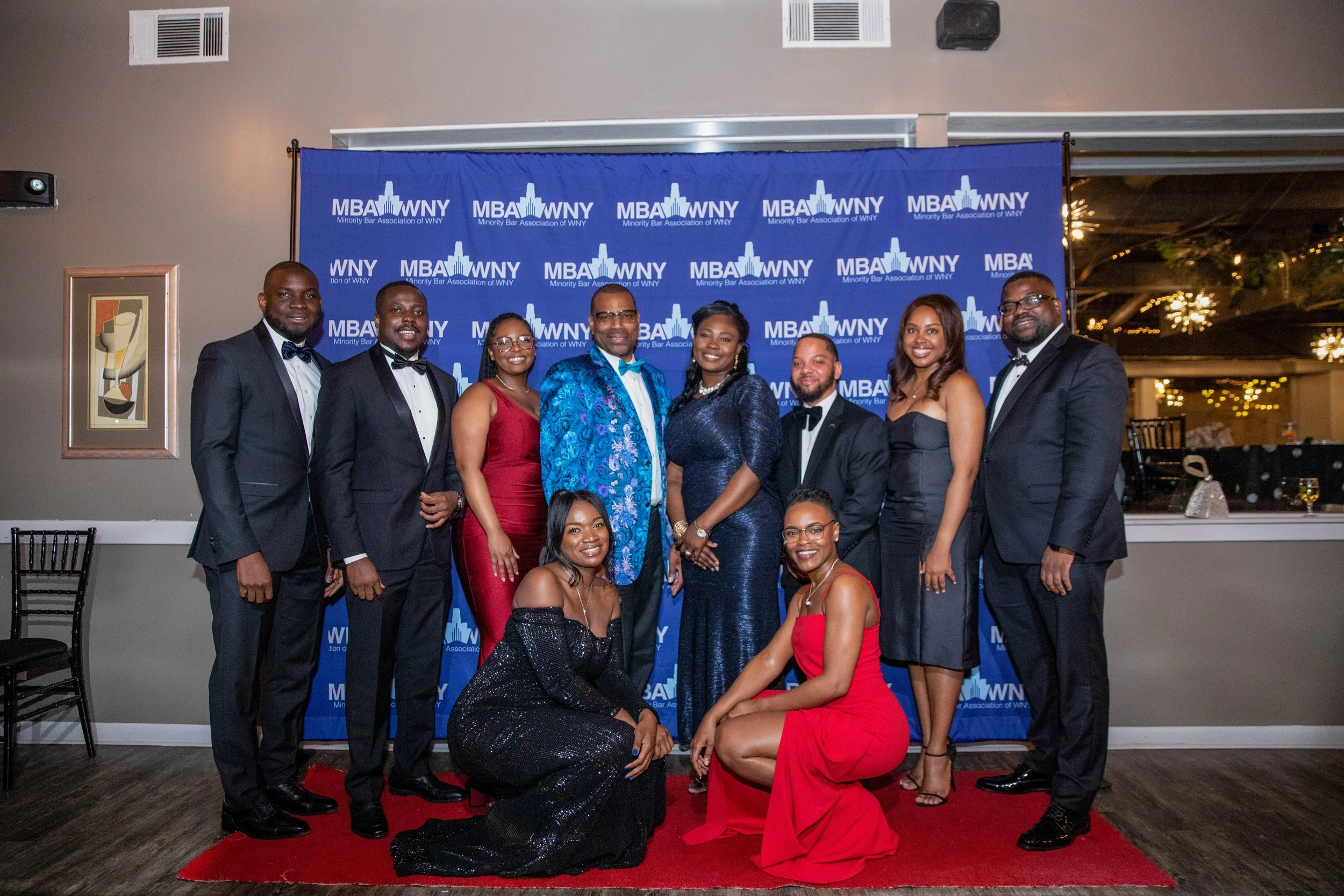 Group of ten professionally dressed people posing for a photo at a formal event with a blue backdrop labeled 'MBA WNY Minority Bar Association of WNY' and colorful lights in the background.