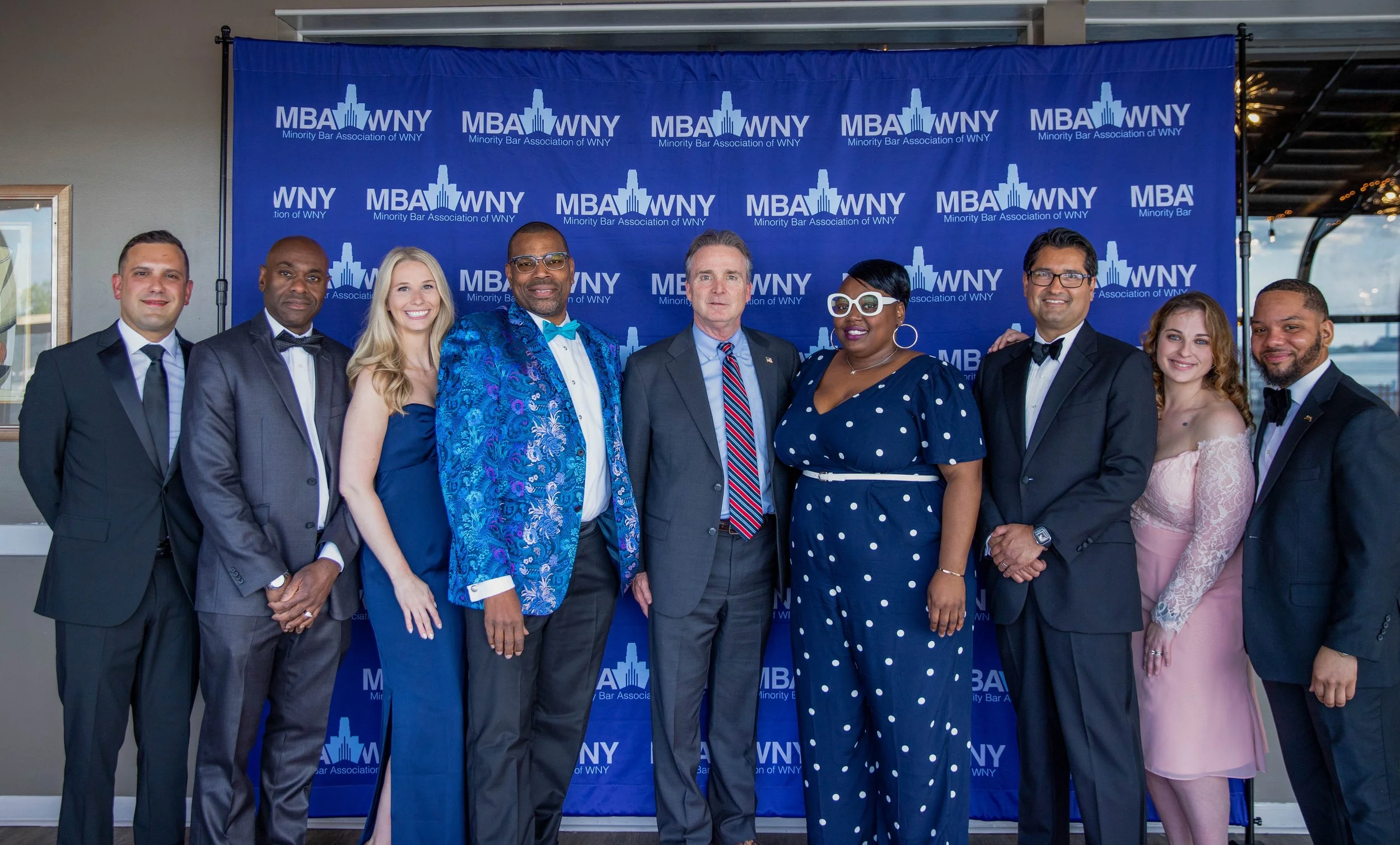 A group of nine diverse people dressed in formal attire, standing in front of a blue backdrop with the logo and text 'MBA WNY Minority Bar Association of WNY.' They are smiling and posing for a photo at an event.