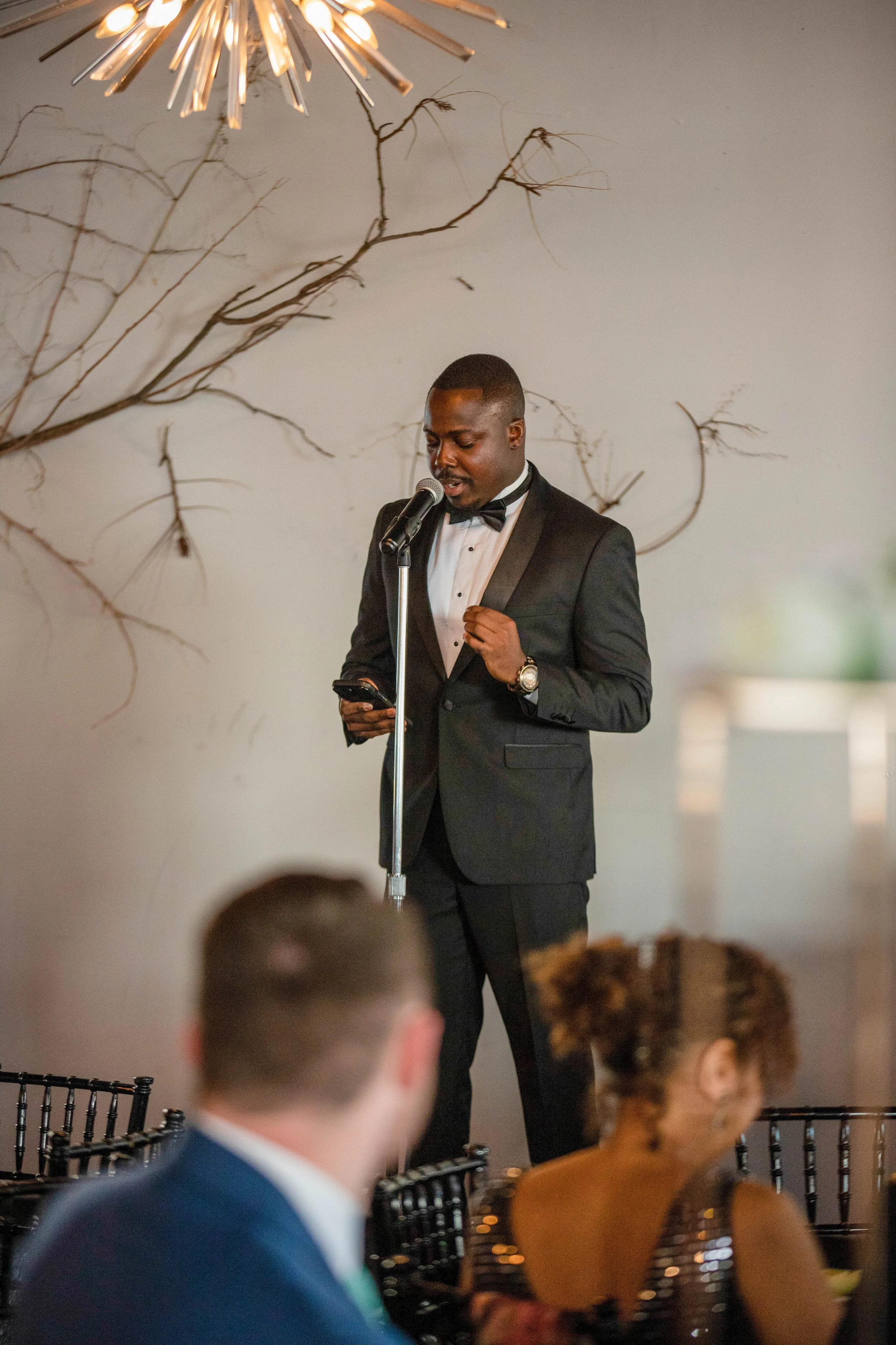 A man in a tuxedo and bowtie is standing at a microphone, reading from his phone. He is in a decorated indoor venue with a rustic branch and modern lamp decor on the wall behind him. Several people are seated in front of him, blurred in the foregroun