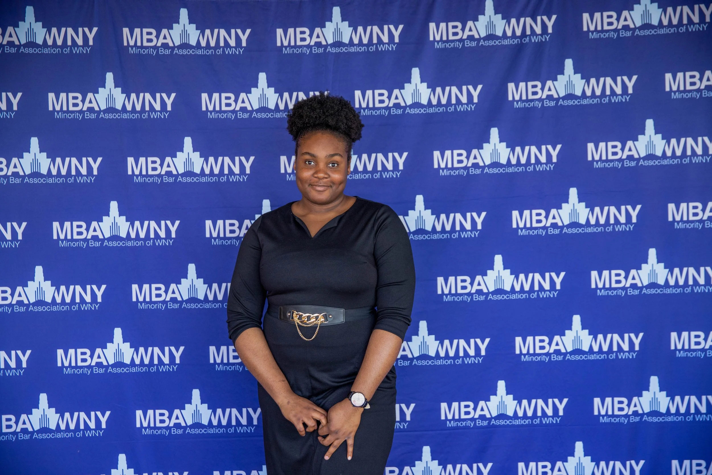 A woman with natural hair styled in a high puff pose standing in front of a blue backdrop with the text 'MBA WNY Minority Bar Association of WNY' repeated in white, smiling, wearing a black top, a belt with a gold chain detail, and a watch.