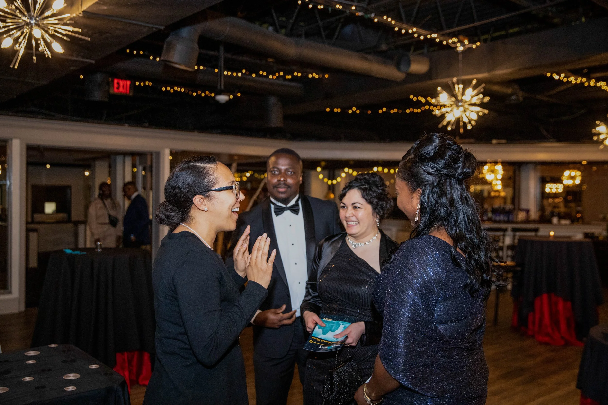 Group of four people at a social event, two women and two men, engaging in conversation. The women are smiling and appear to be enjoying each other's company, with one woman holding a brochure. The man in the background is dressed in a tuxedo.