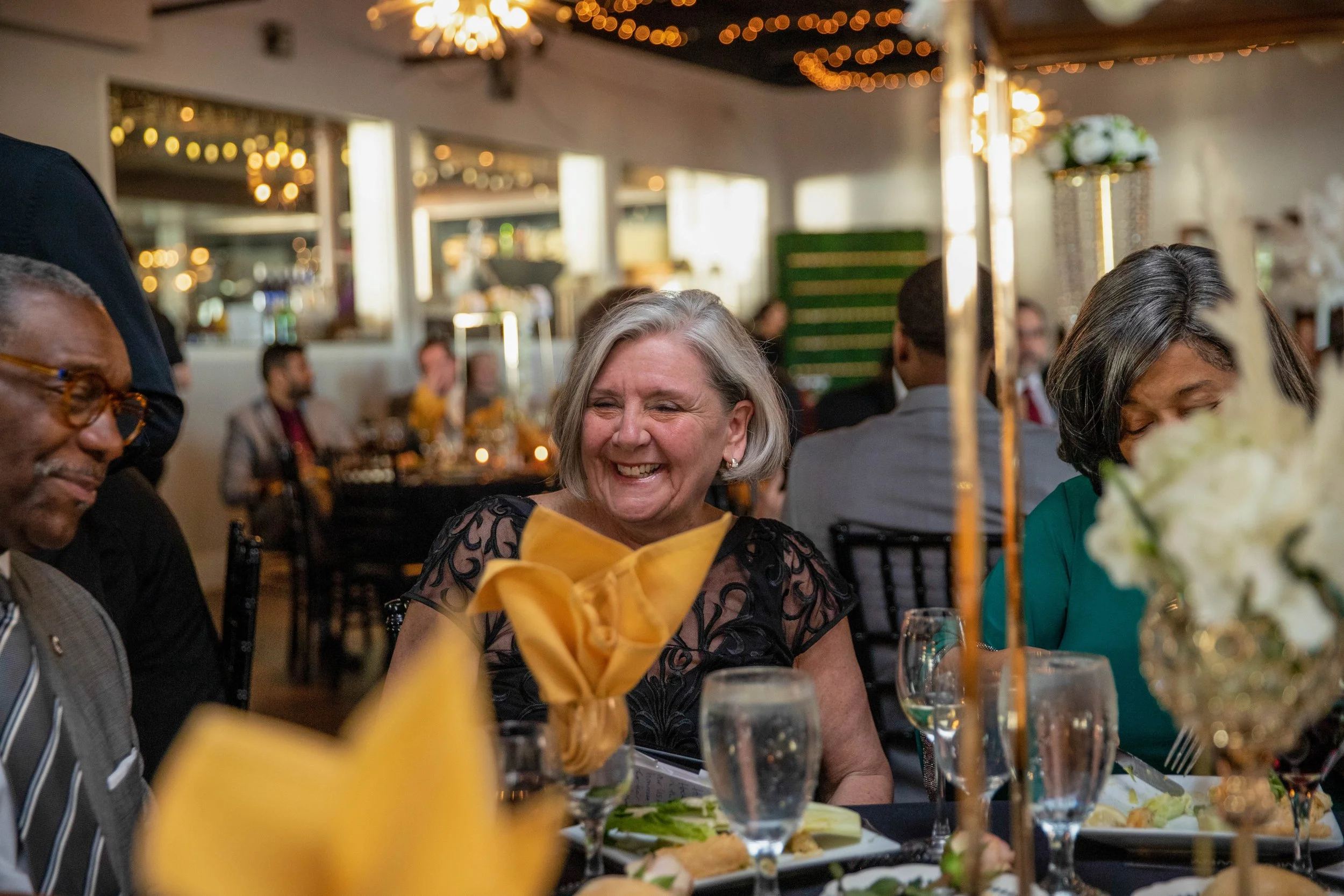 People attending a celebration or dinner party in a decorated indoor venue, with tables set with food, drinks, and floral centerpieces, smiling and engaging with each other.