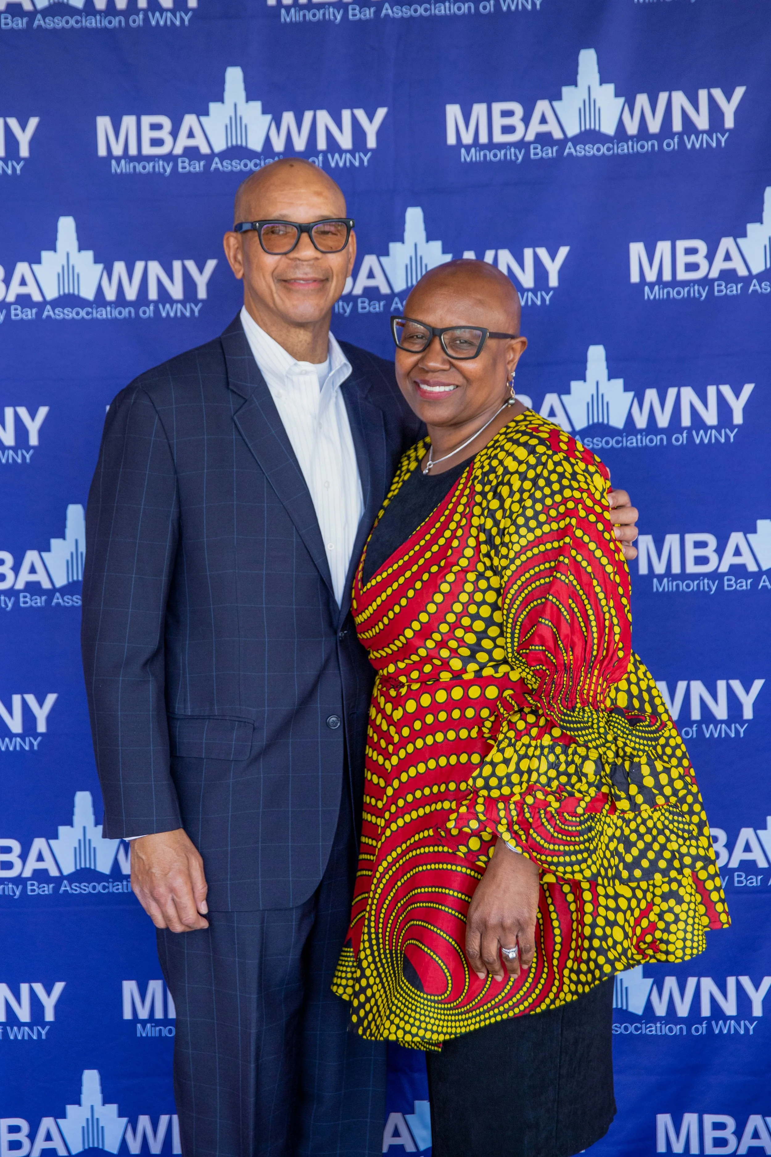 Two people, a man and a woman, standing in front of a blue backdrop with the text 'MBA WNY Minority Bar Association of WNY.' The man is wearing a dark checkered suit, white shirt, glasses, and is smiling. The woman is wearing a vibrant red, yellow, a