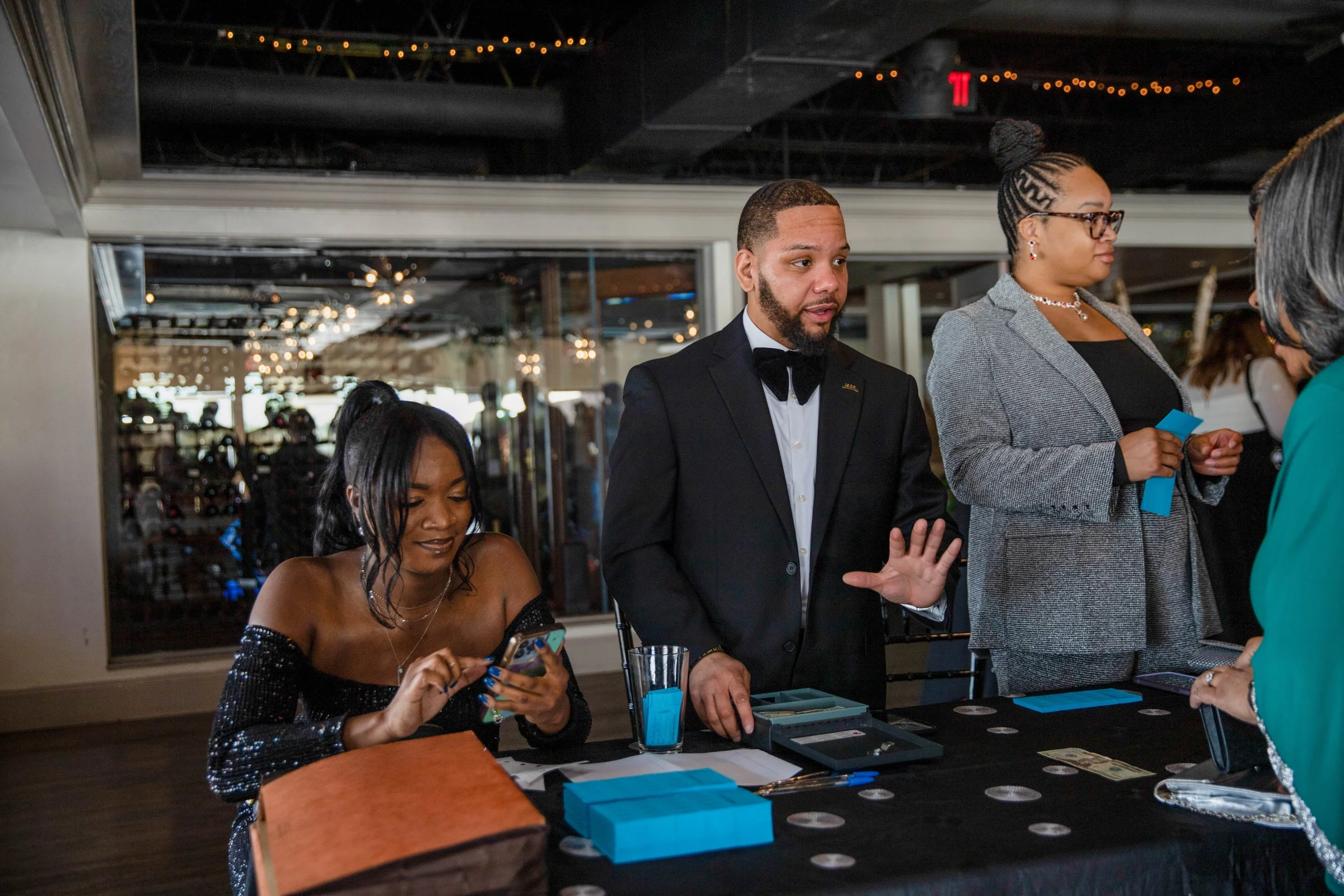People in formal attire at a crowded event, engaged in conversation and using their phones, with a table displaying money, cards, and blue boxes.