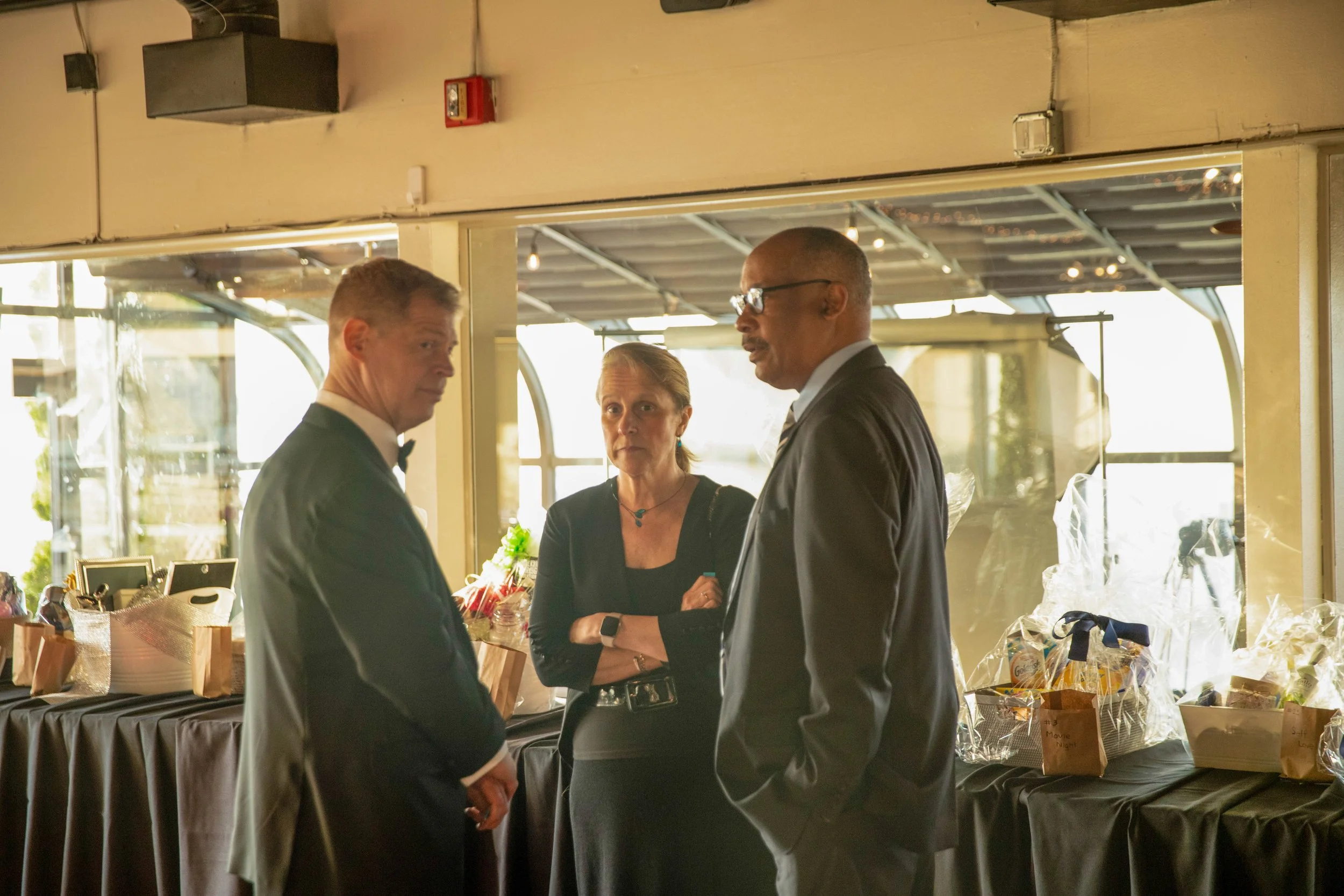 Three people dressed in formal attire are engaged in conversation inside a well-lit room with large windows. The background features a table with gift bags and a tablecloth, possibly at a social event or gathering.