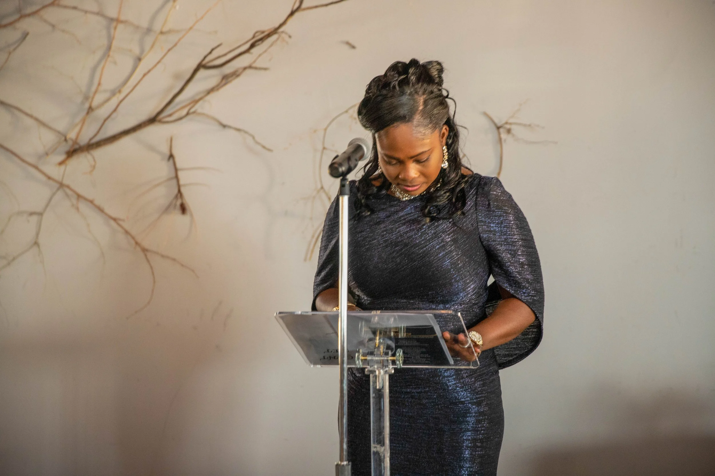 A woman dressed in a shiny black dress standing behind a clear podium, looking down at a paper or notes, with a microphone in front of her, against a light-colored wall with dried branches for decoration.