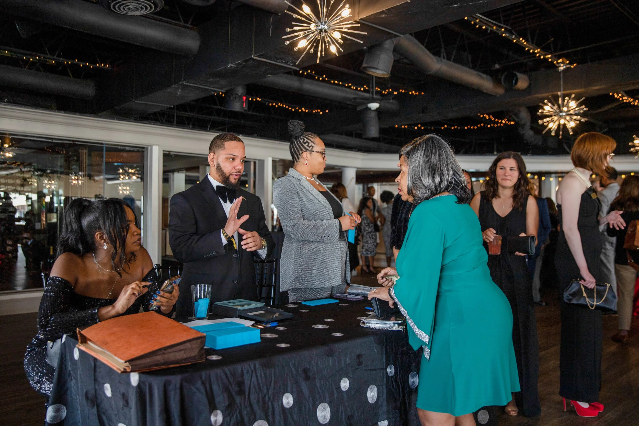 People gather at a registration or check-in table during an indoor event, with decorations and warm lighting overhead.