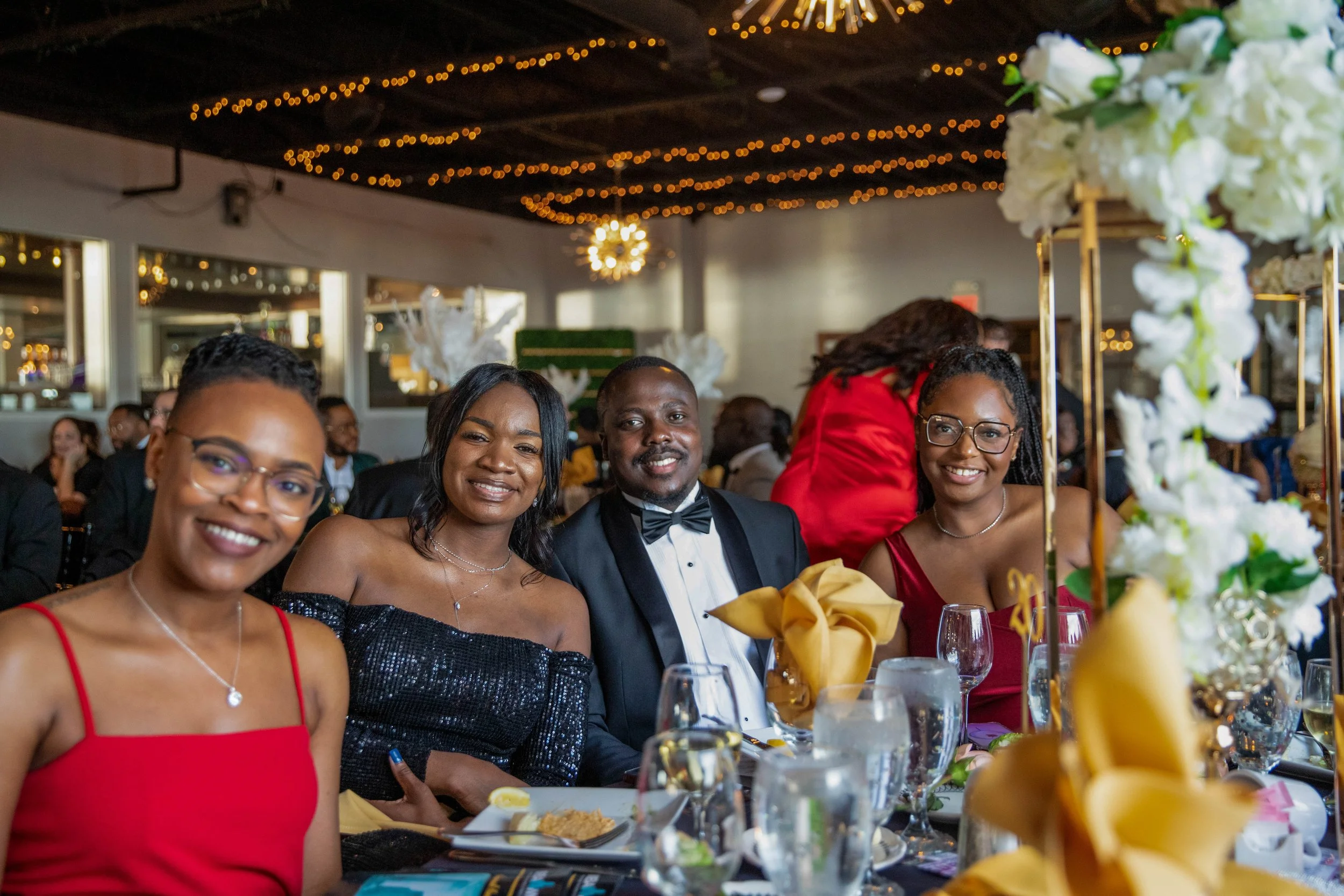 A group of people dressed formally, including women in dresses and a man in a tuxedo, sitting at a decorated banquet table with flowers and glasses, at a festive event with string lights and a ceiling decoration.