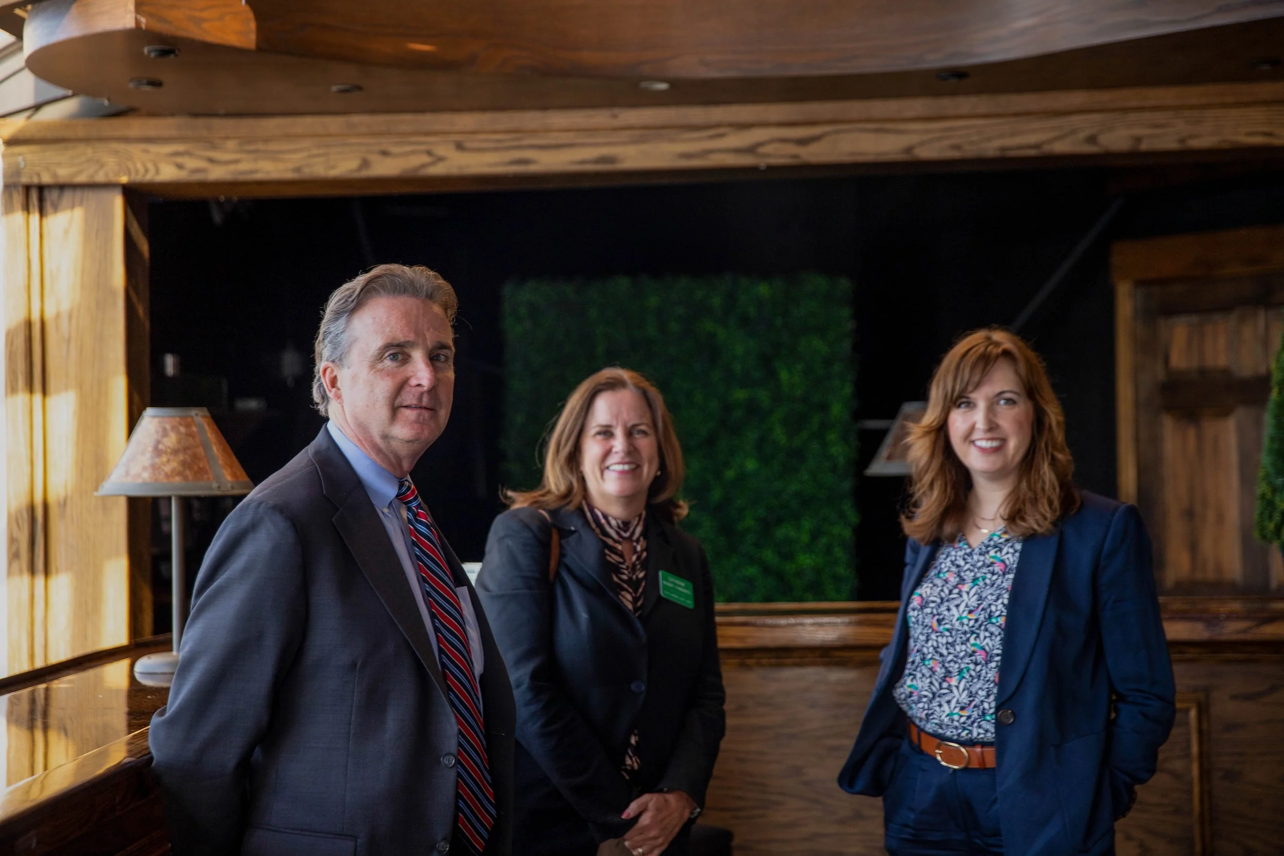 Four professionally dressed individuals standing and smiling inside a warm, wood-paneled room with green decorative wall features. The group includes two women and two men, with the man on the left wearing a dark suit and striped tie.
