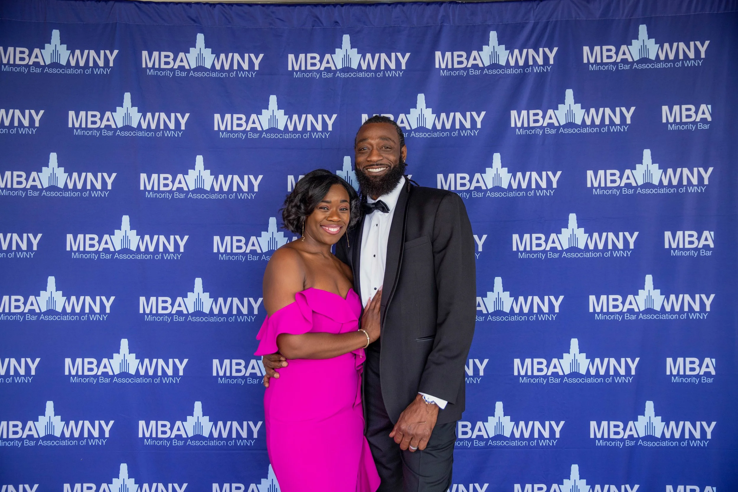 A man and woman dressed formally, standing in front of a blue backdrop with the logo and text "MBA WNY Minority Bar Association of WNY." The woman is wearing a strapless pink dress, and the man is in a black tuxedo with a bow tie. They are smiling an