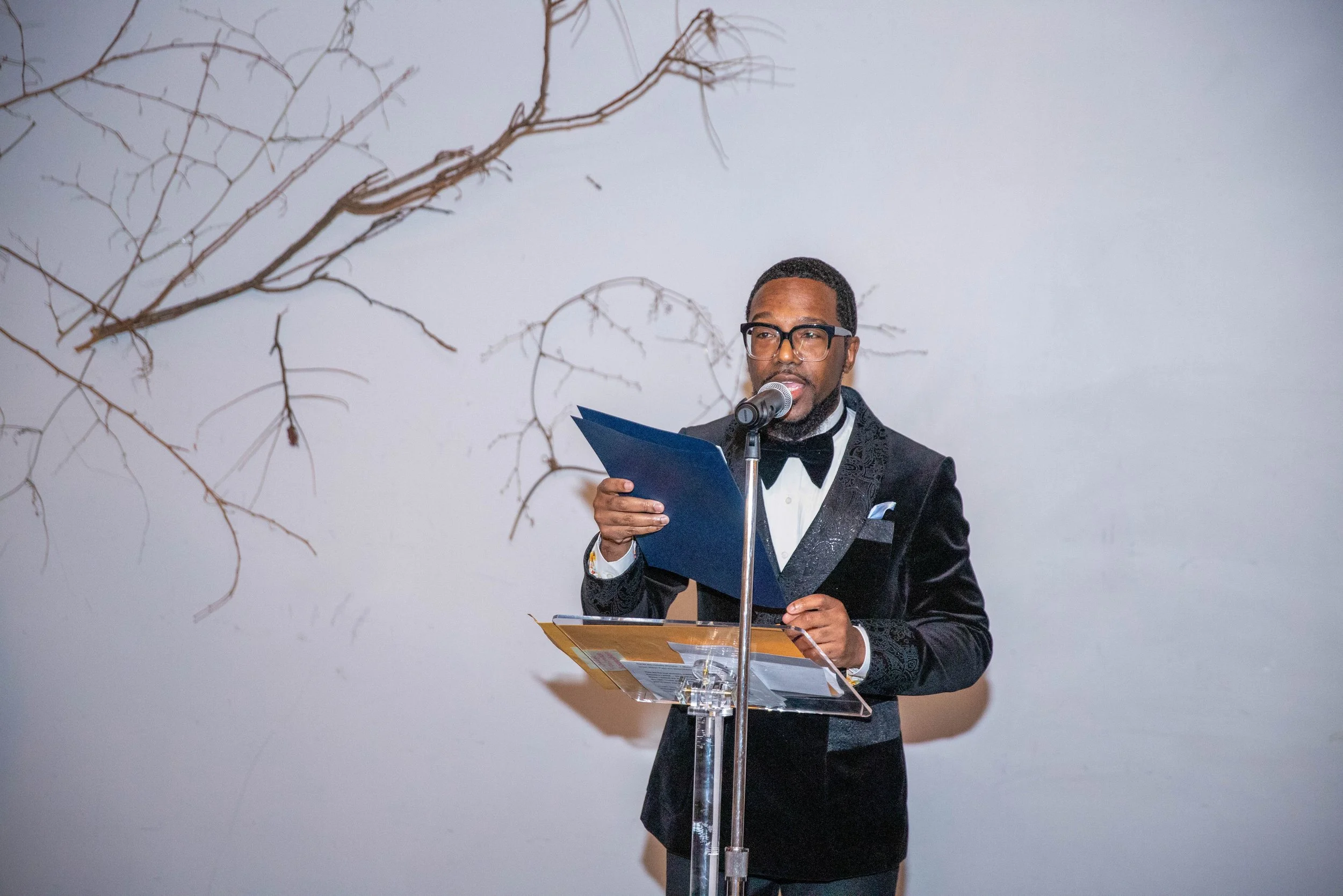 A man dressed formally in a tuxedo with a bowtie, wearing glasses, is standing at a clear podium. He is reading from a blue folder into a microphone. Behind him, there is a plain white wall with some leafless branches or twigs mounted on it.