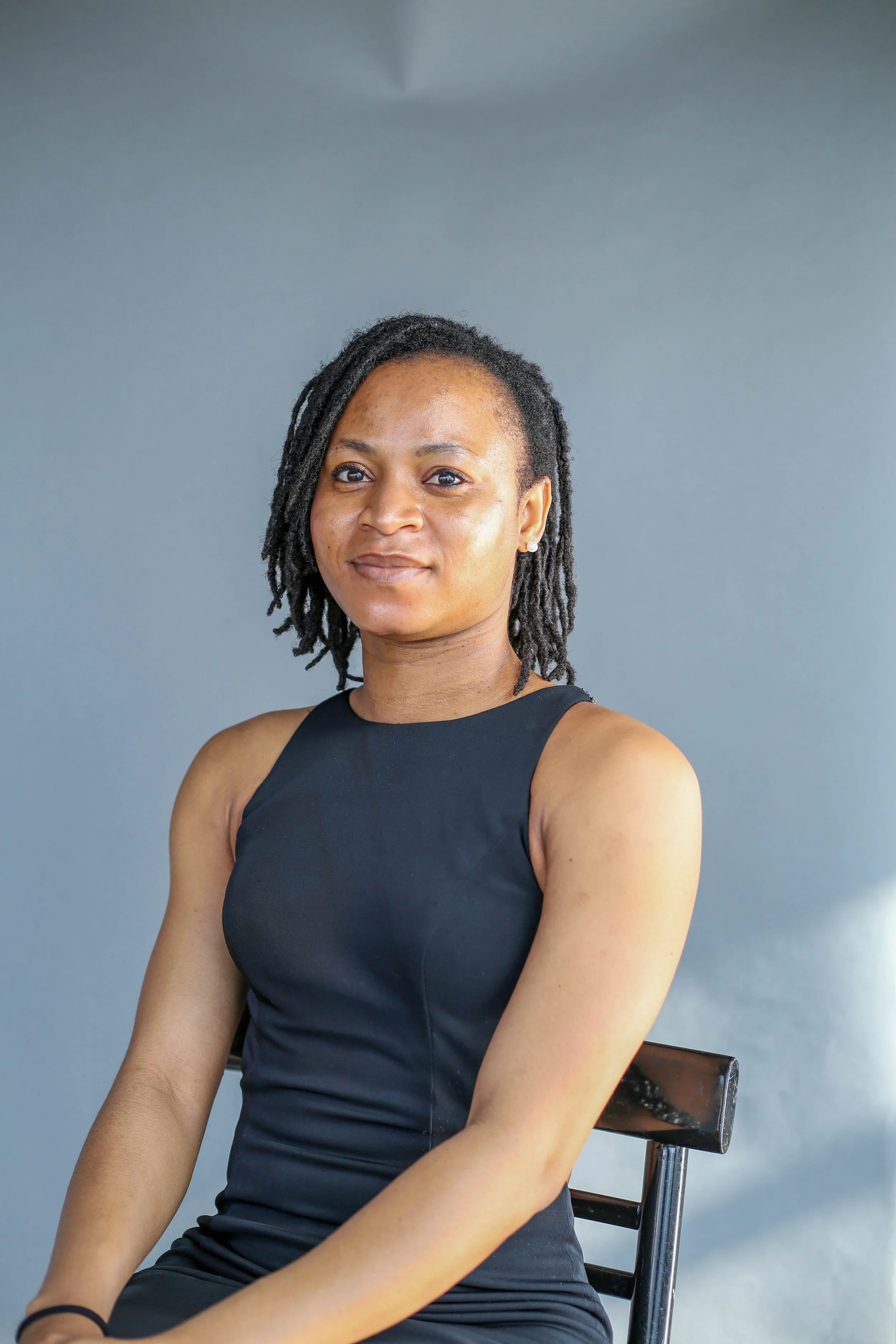 A woman with shoulder-length dreadlocks wearing a black sleeveless top, sitting on a black chair against a gray background.