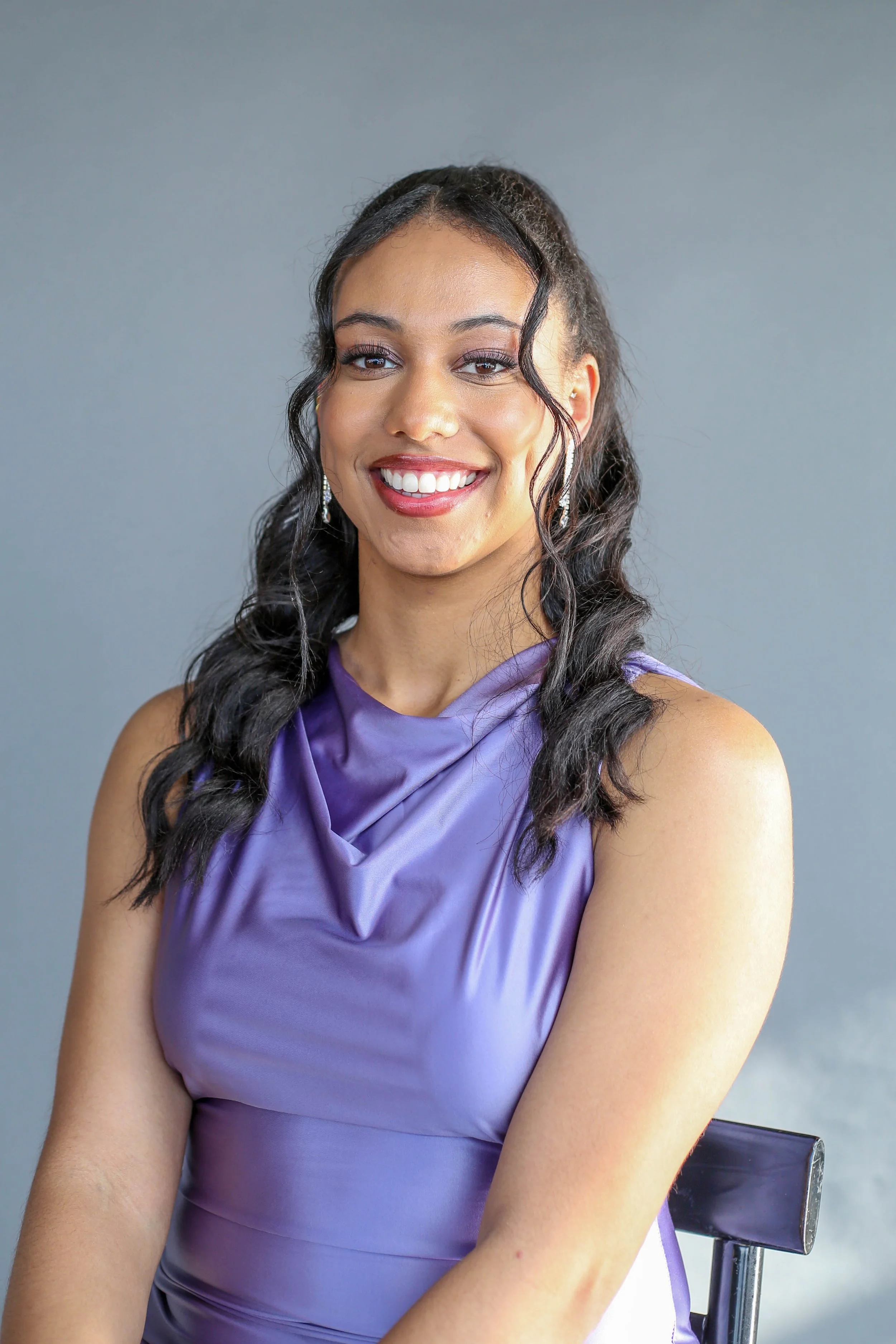 A woman with long, dark, curly hair styled in a high ponytail, wearing a purple satin dress with a draped neckline, and silver earrings, smiling while sitting on a black chair with a gray background.