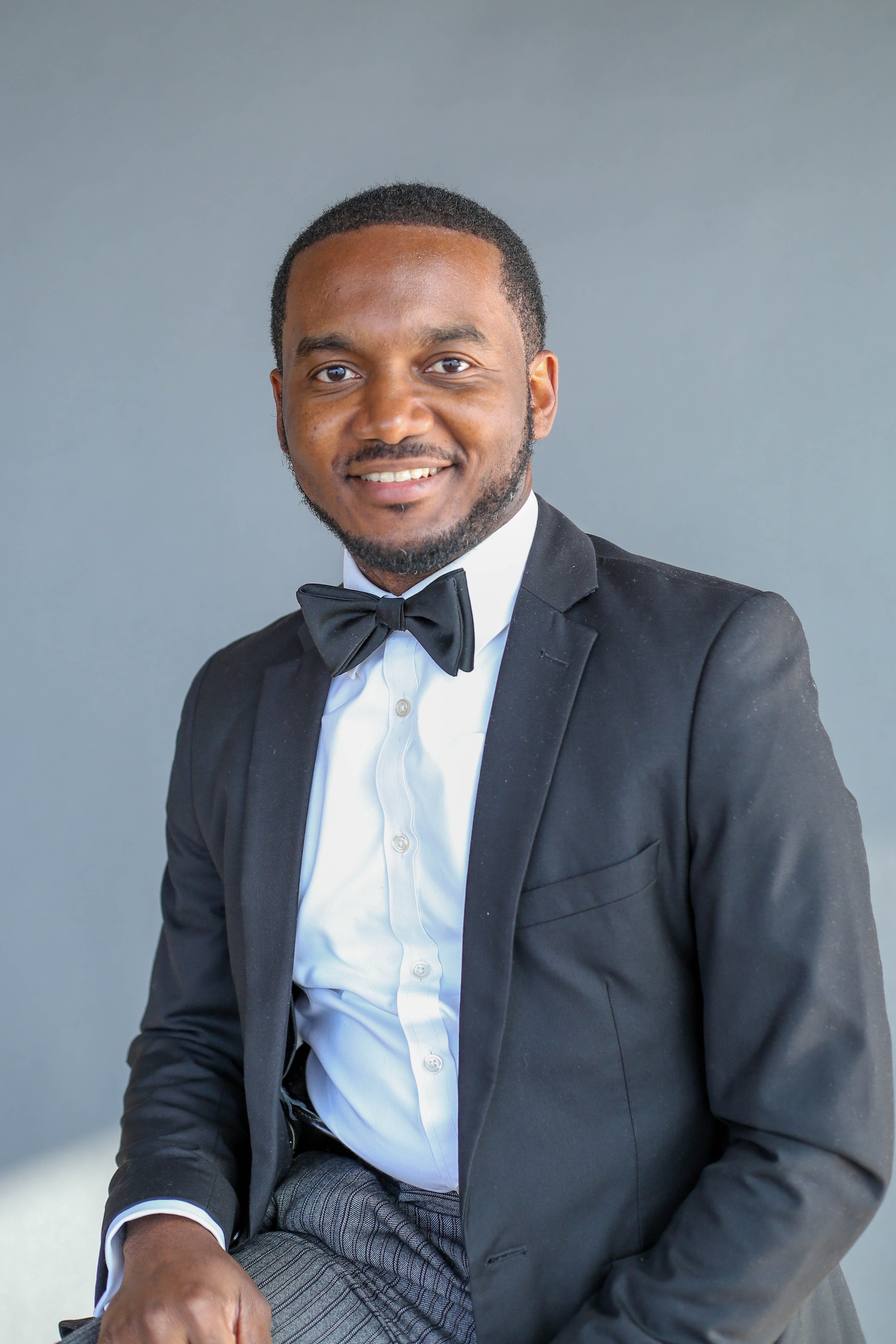 Portrait of a smiling man wearing a black tuxedo and bowtie, seated against a plain gray background.