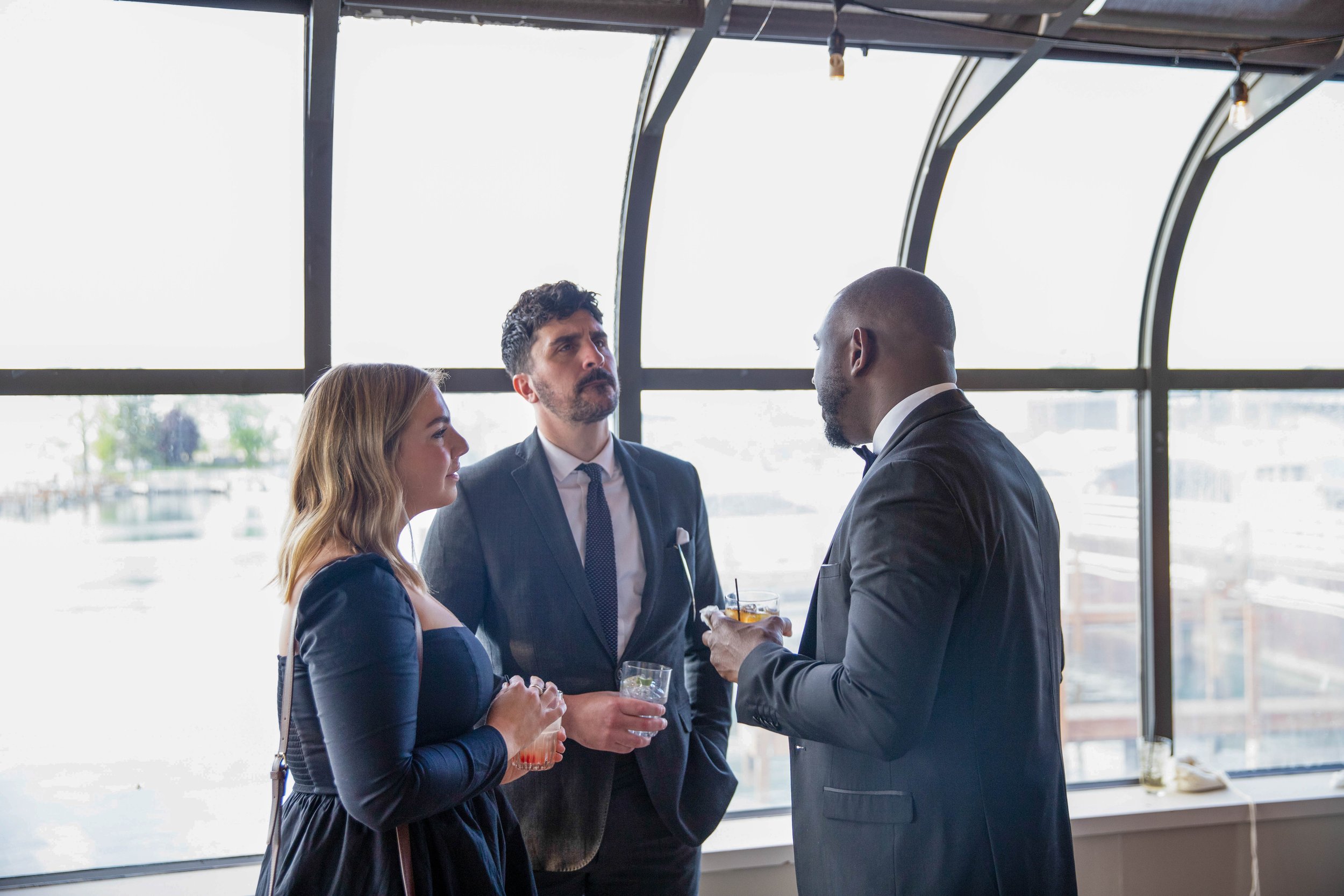 Three people in formal attire having a conversation at an indoor event with large windows in the background.