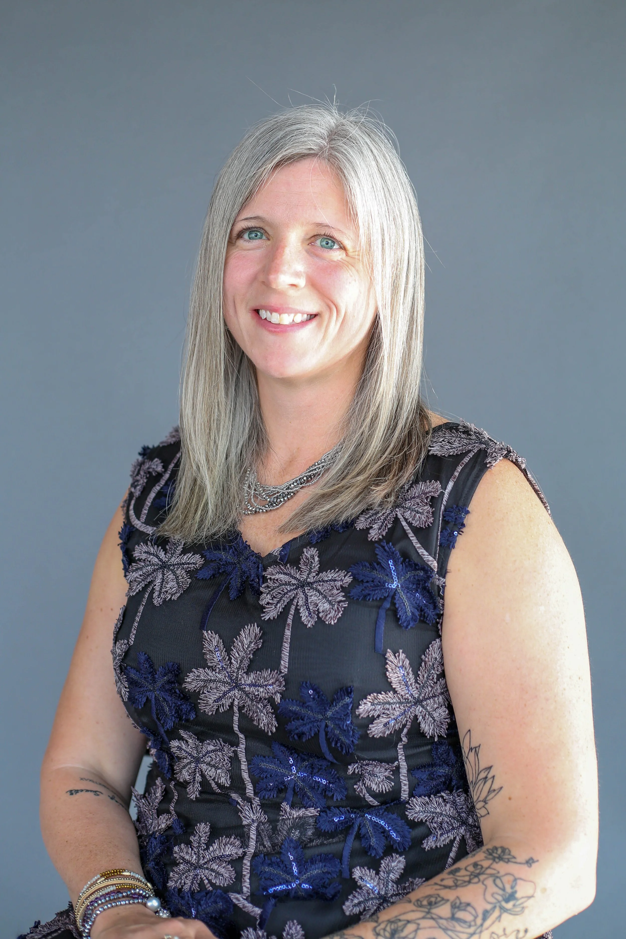 A middle-aged woman with long silver hair and blue eyes, smiling and wearing a sleeveless black dress with purple and silver floral embroidery, silver jewelry, and tattoos on her arms, posed against a plain gray background.