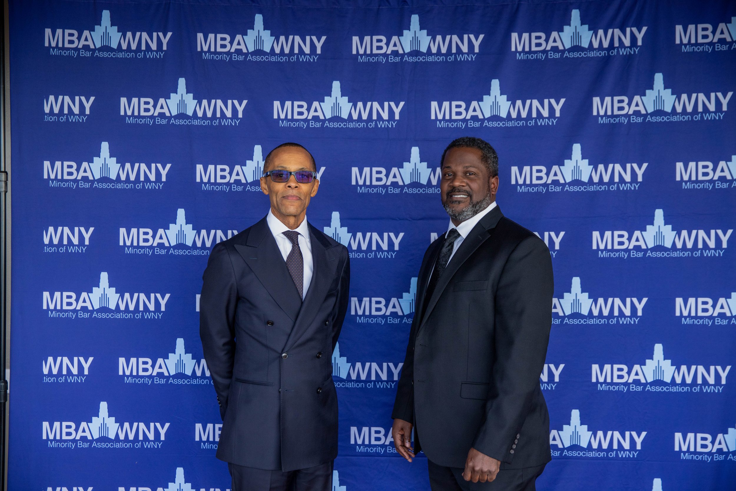 Two men in suits standing in front of a blue backdrop with the logo of the Minority Bar Association of WNY, featuring a silhouette of a city skyline and the text 'MBA WNY Minority Bar Association of WNY' repeated across the background.