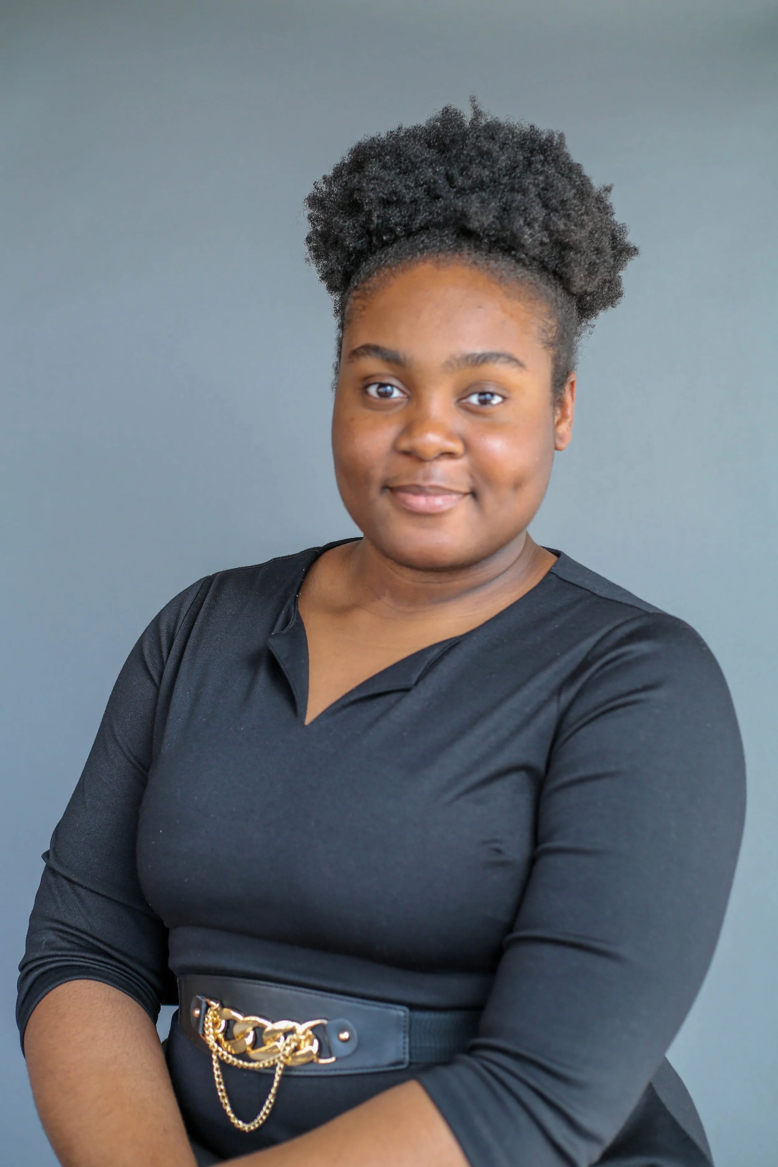 A woman with dark skin and natural curly hair styled in an updo, wearing a black top and a black belt with gold chain details, posing against a plain gray background.