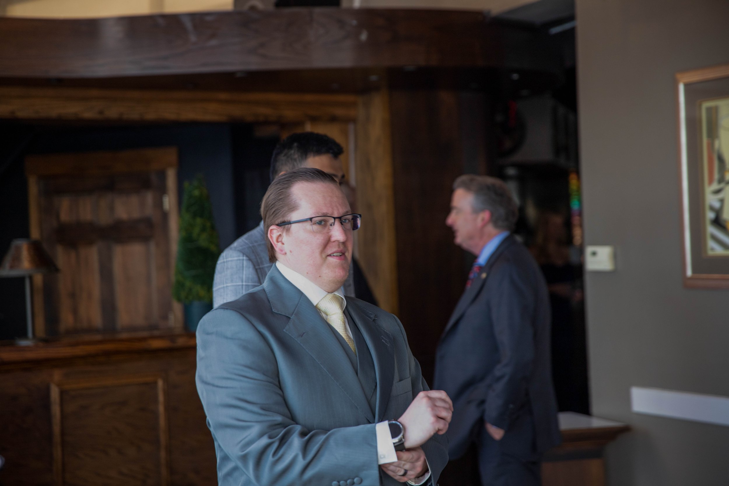 A man in a blue suit and yellow tie with glasses standing indoors, shaking hands with another man in a suit.