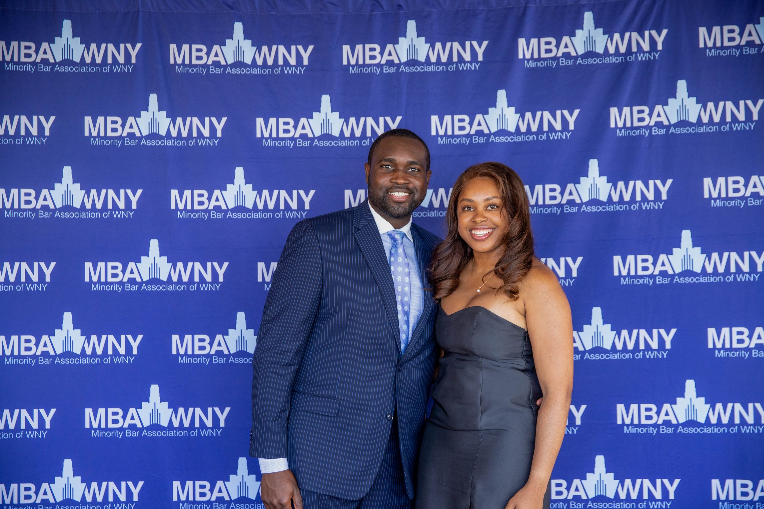 A man and woman dressed in formal attire smiling, standing in front of a blue backdrop with the logo and text for the Minority Bar Association of WNY.