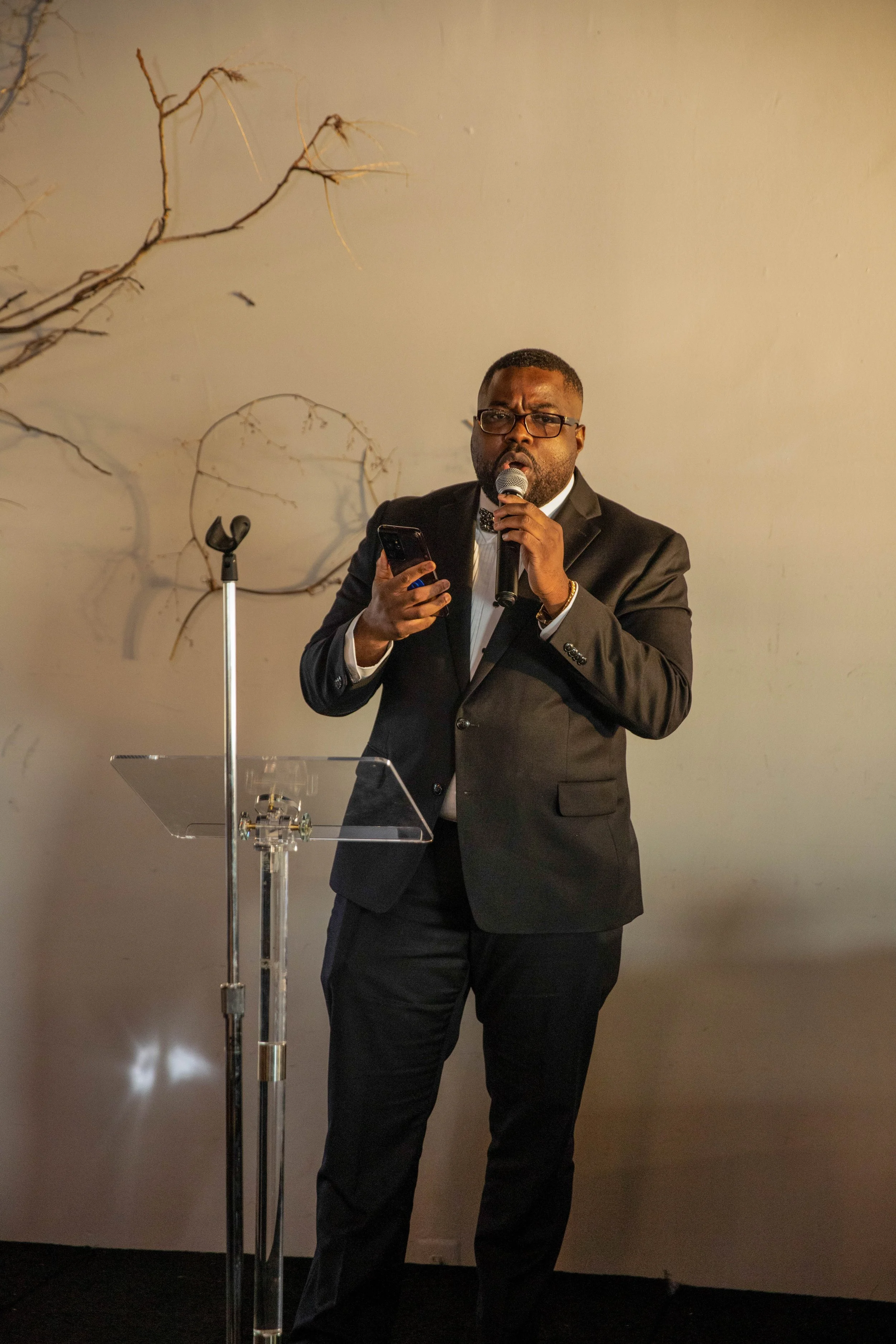 A man wearing a black suit, white shirt, and black bow tie stands at a clear podium, holding a microphone in one hand and a phone in the other. He is speaking into the microphone with a serious expression. There is a microphone stand beside him, and 