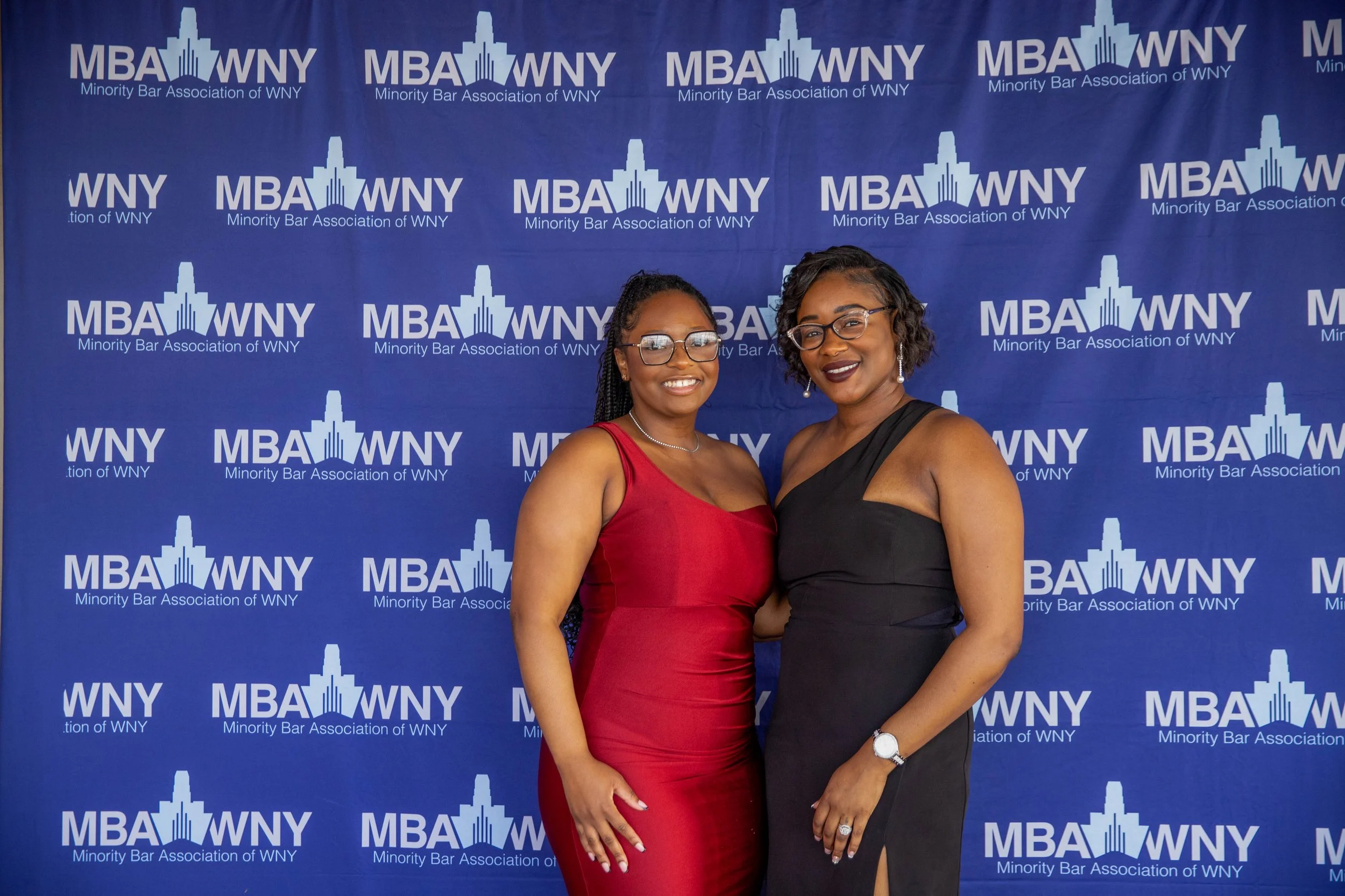 Two women in formal dresses are standing in front of a blue backdrop with the logo and text for the Minority Bar Association of WNY. One woman is wearing a red dress and glasses, and the other is wearing a black dress and glasses. They are smiling an
