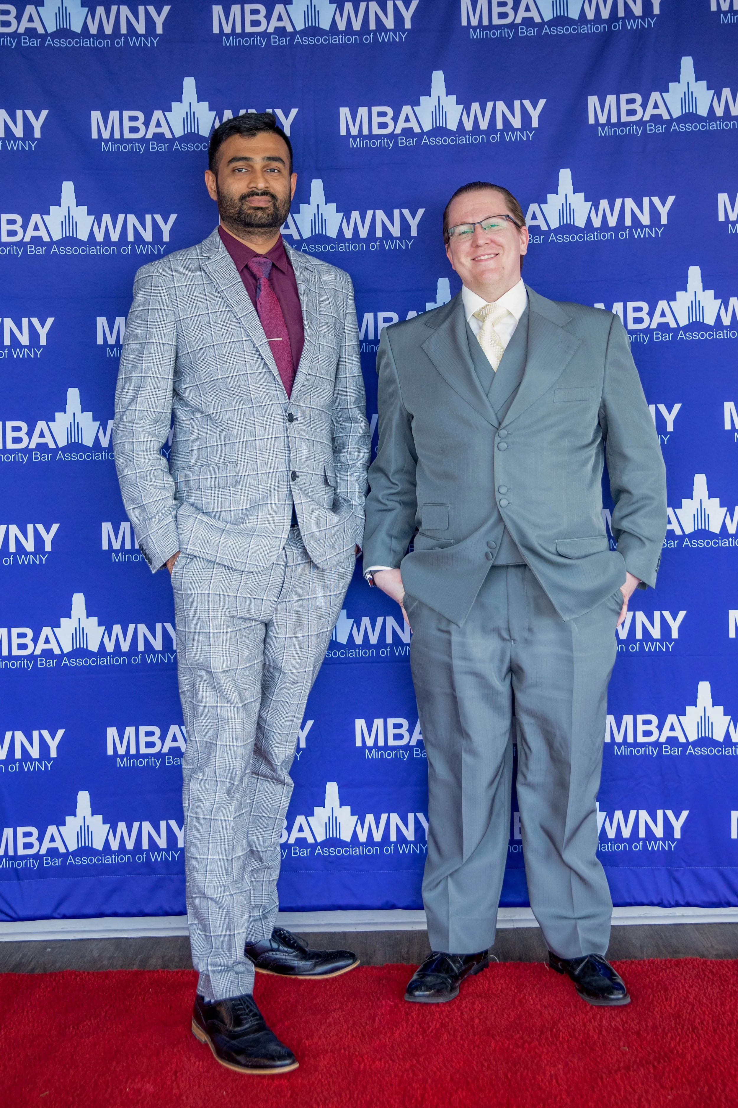 Two men dressed in suits standing in front of a blue backdrop with logos of the Minority Bar Association of WNY. One man has darker skin and is wearing a patterned gray suit with a maroon shirt and tie, the other has lighter skin, glasses, and is wea