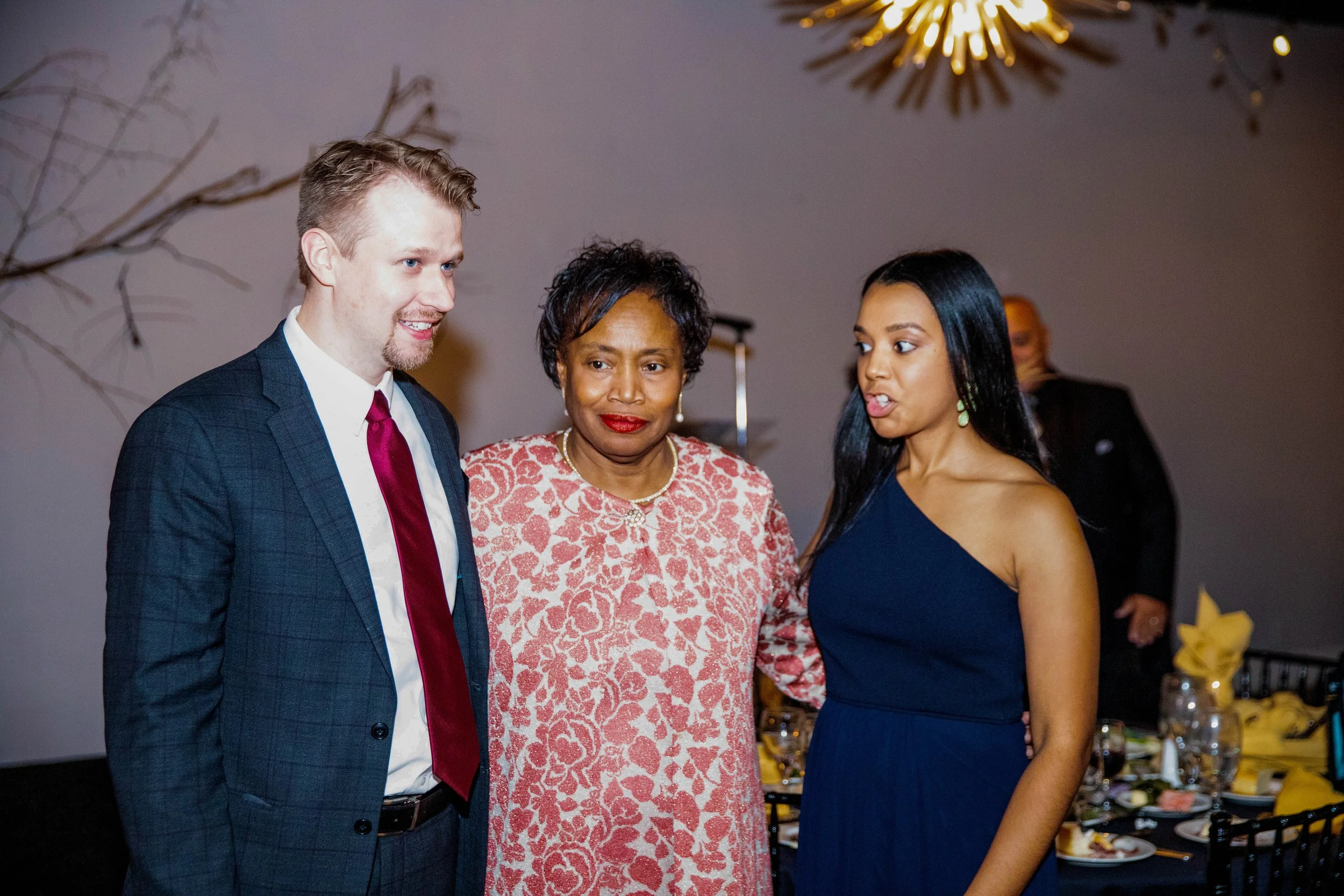 Three people, a man and two women, are standing together at an indoor event, engaged in conversation. The man is wearing a dark suit with a white shirt and red tie, the woman in the middle has short curly hair and is wearing a pink and white patterne