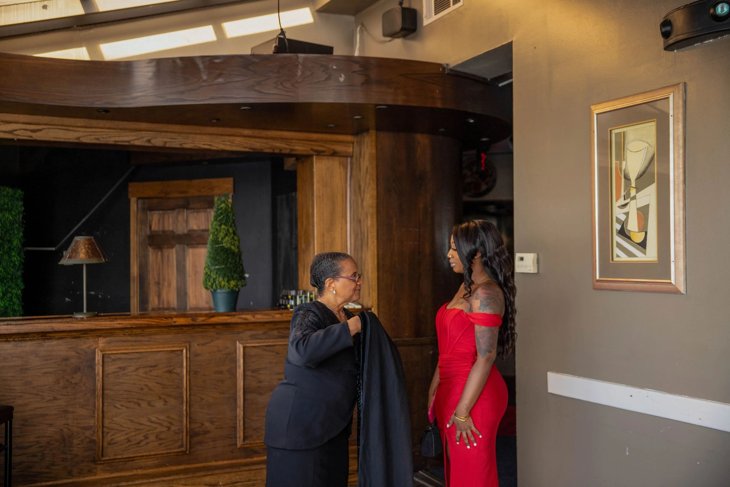 Two women are standing and talking to each other in a restaurant or bar, one is wearing a black jacket and glasses, and the other is wearing a red dress with tattoos on her arm.