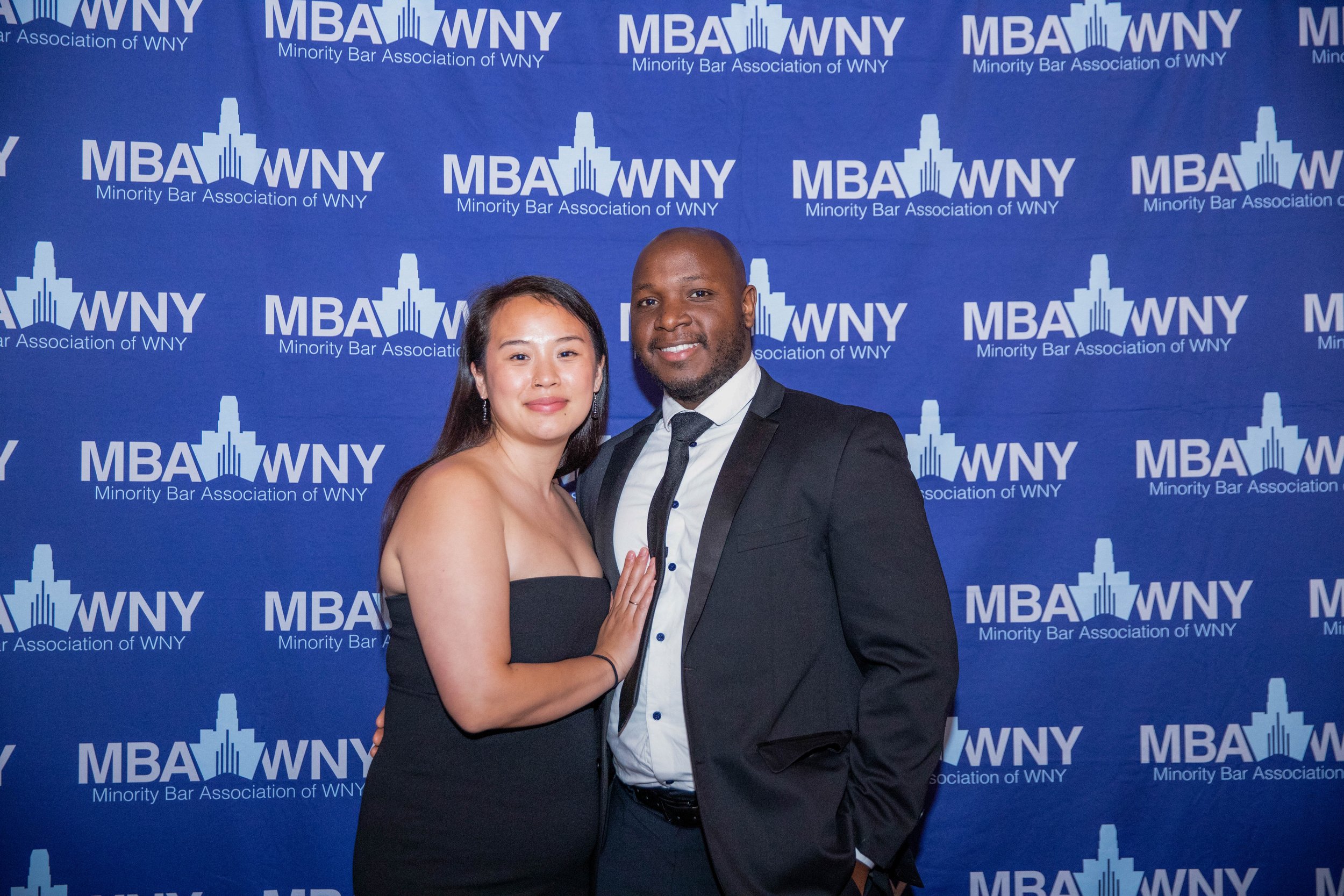 Two people dressed in formal attire, a woman and a man, standing together in front of a blue backdrop with the logo of the Minority Bar Association of WNY and the text. The woman is wearing a black strapless dress and earrings, and the man is wearing