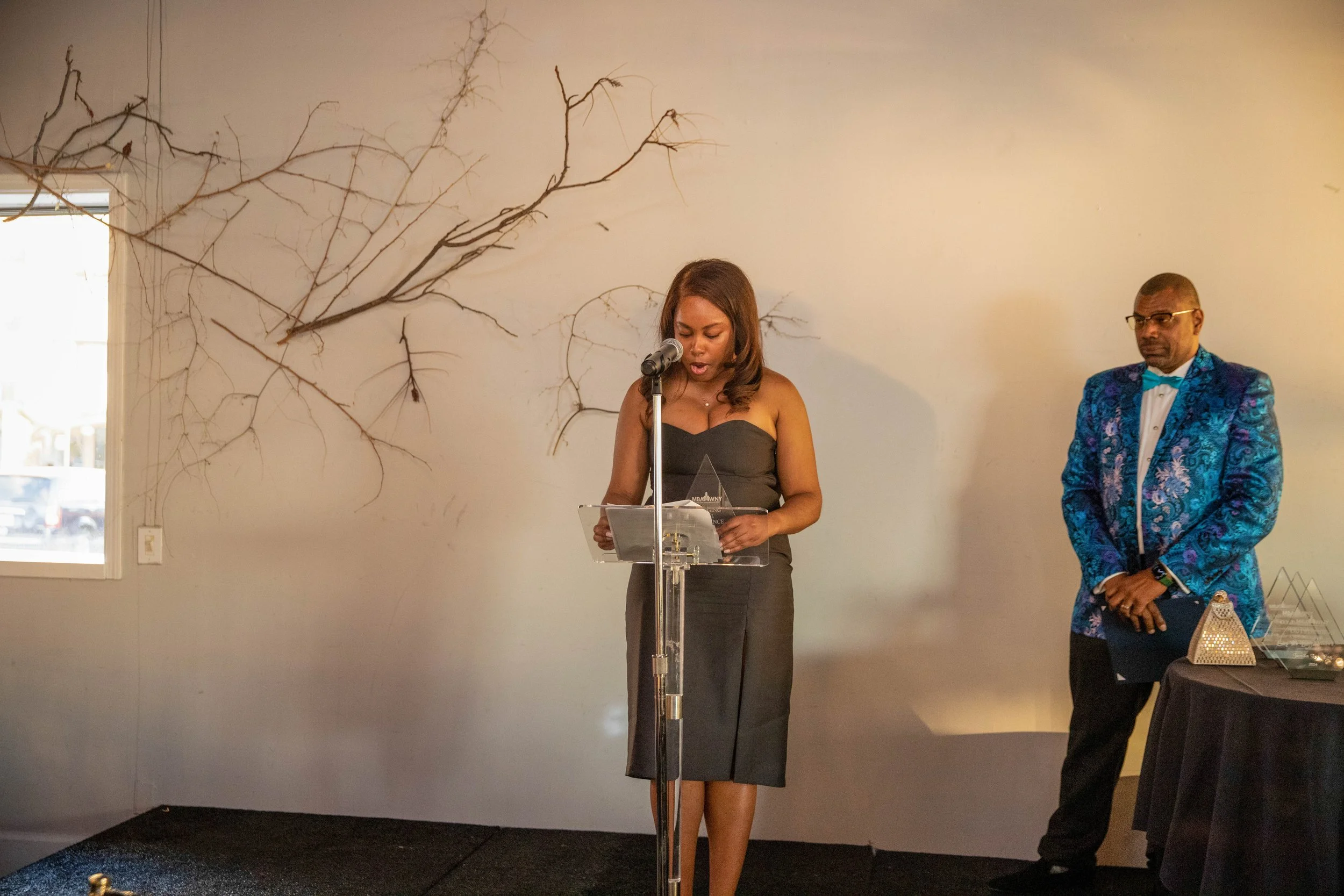 Woman in a black strapless dress speaking at a podium during an event, with a man in a colorful blue jacket standing to her right, against a white wall with decorative branches.