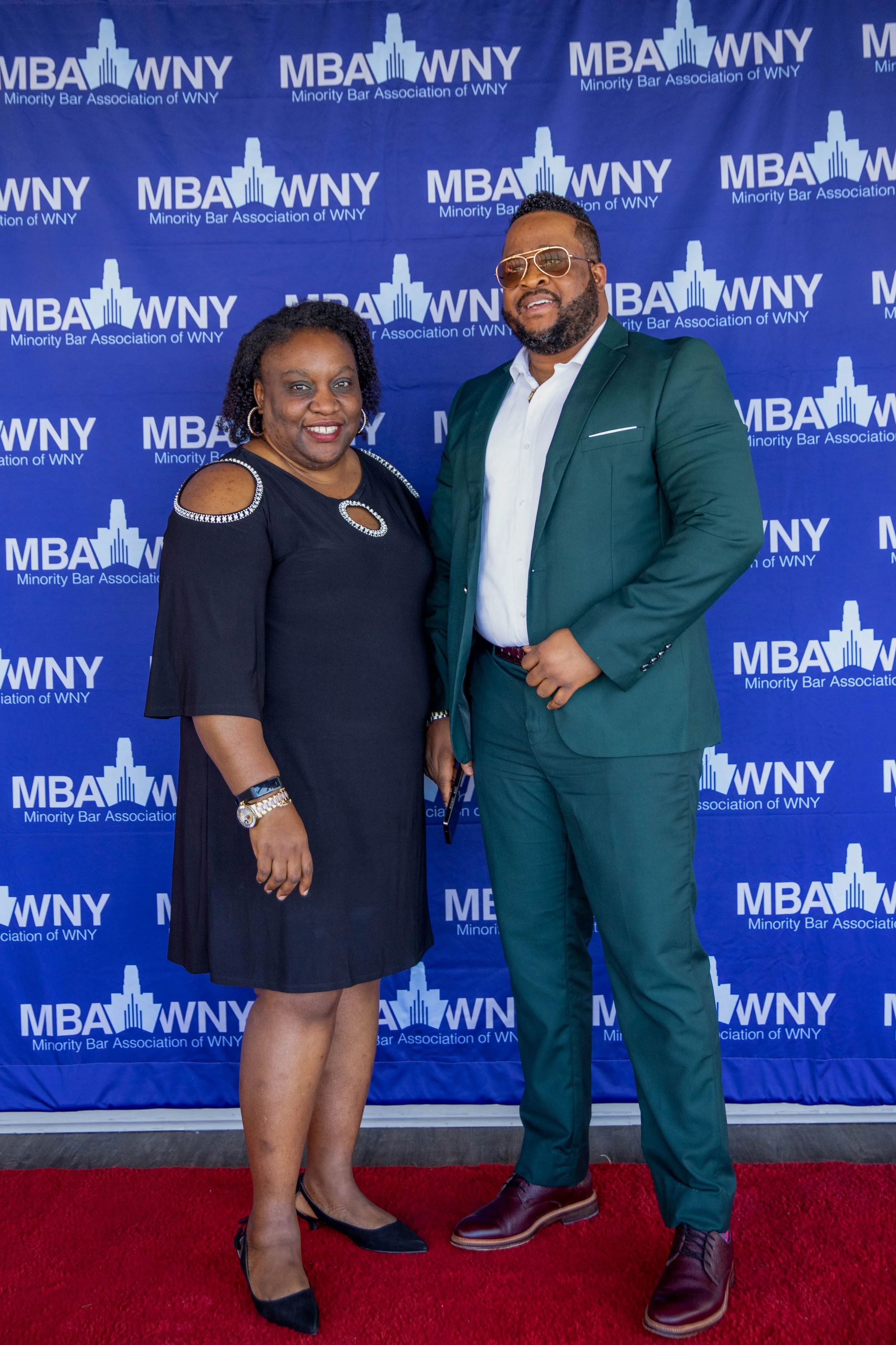 Two people standing on a red carpet beneath a blue backdrop with the logo of the Minority Bar Association of WNY, smiling and posing for a photo. The woman on the left is wearing a black dress with cut-out shoulders and black heels, and the man on th