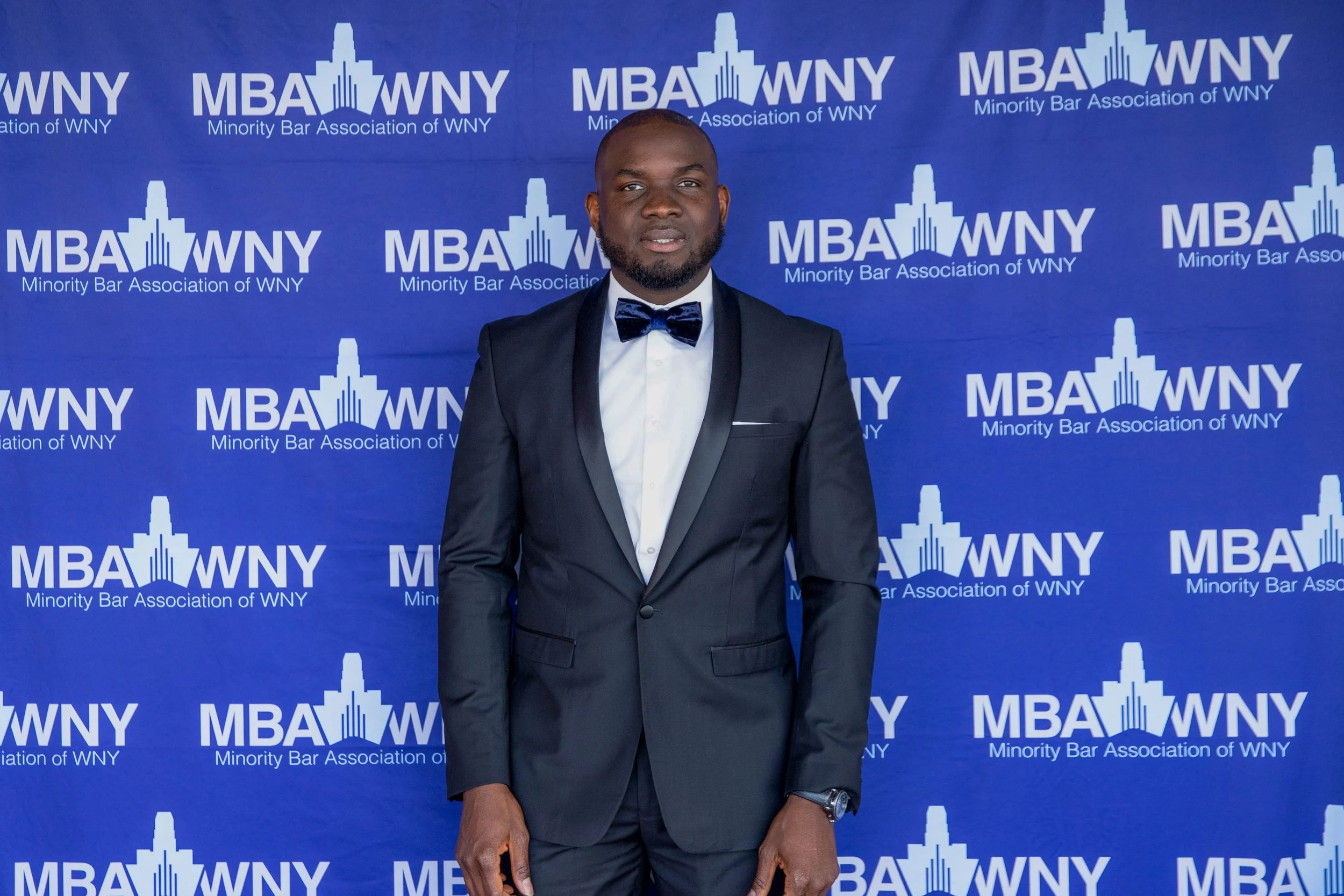 Man in a tuxedo with a bow tie standing in front of a blue backdrop with the logo of the Minority Bar Association of WNY.