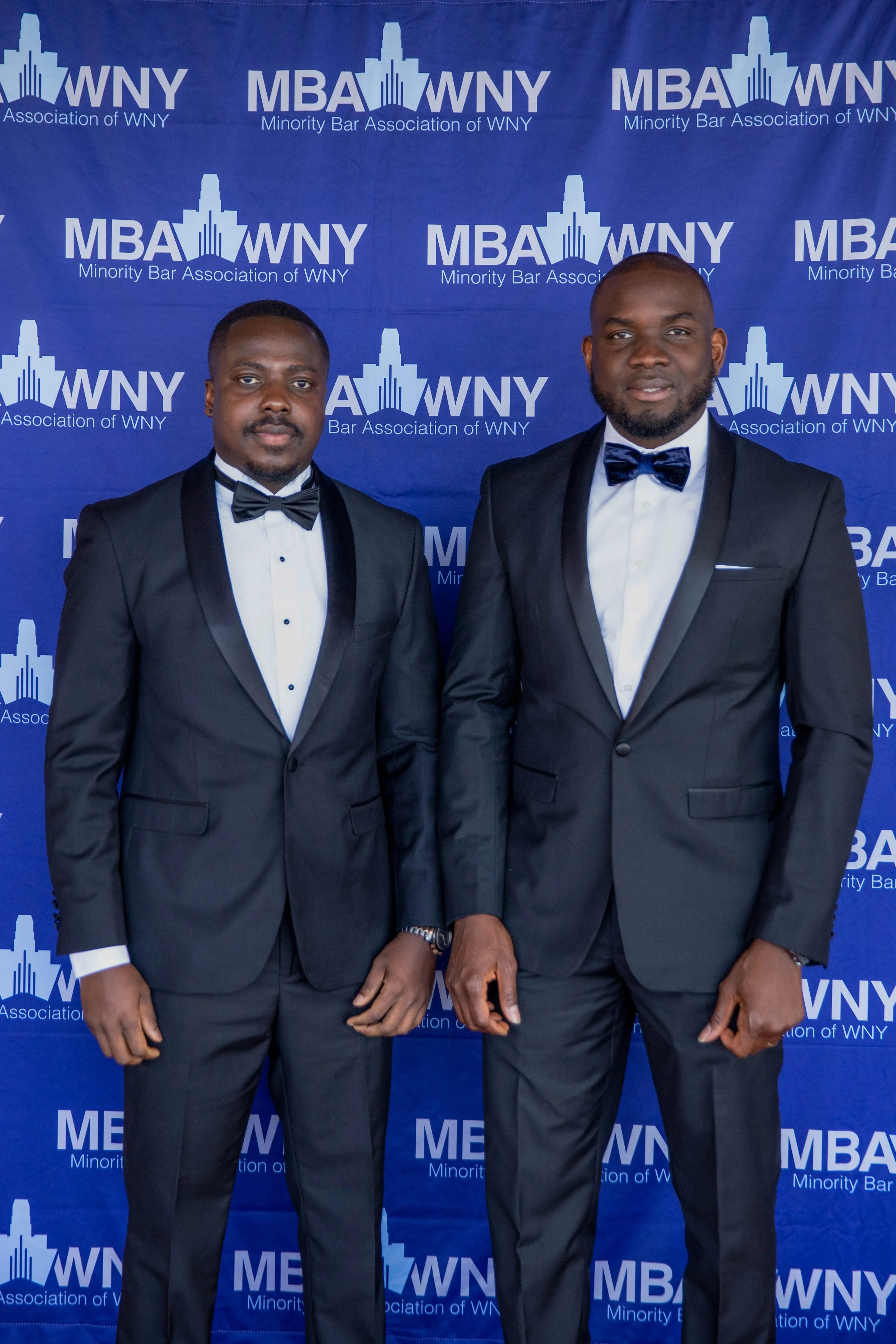 Two men in formal tuxedos posing in front of a blue backdrop with logos reading "MBA WNY Minority Bar Association of WNY."