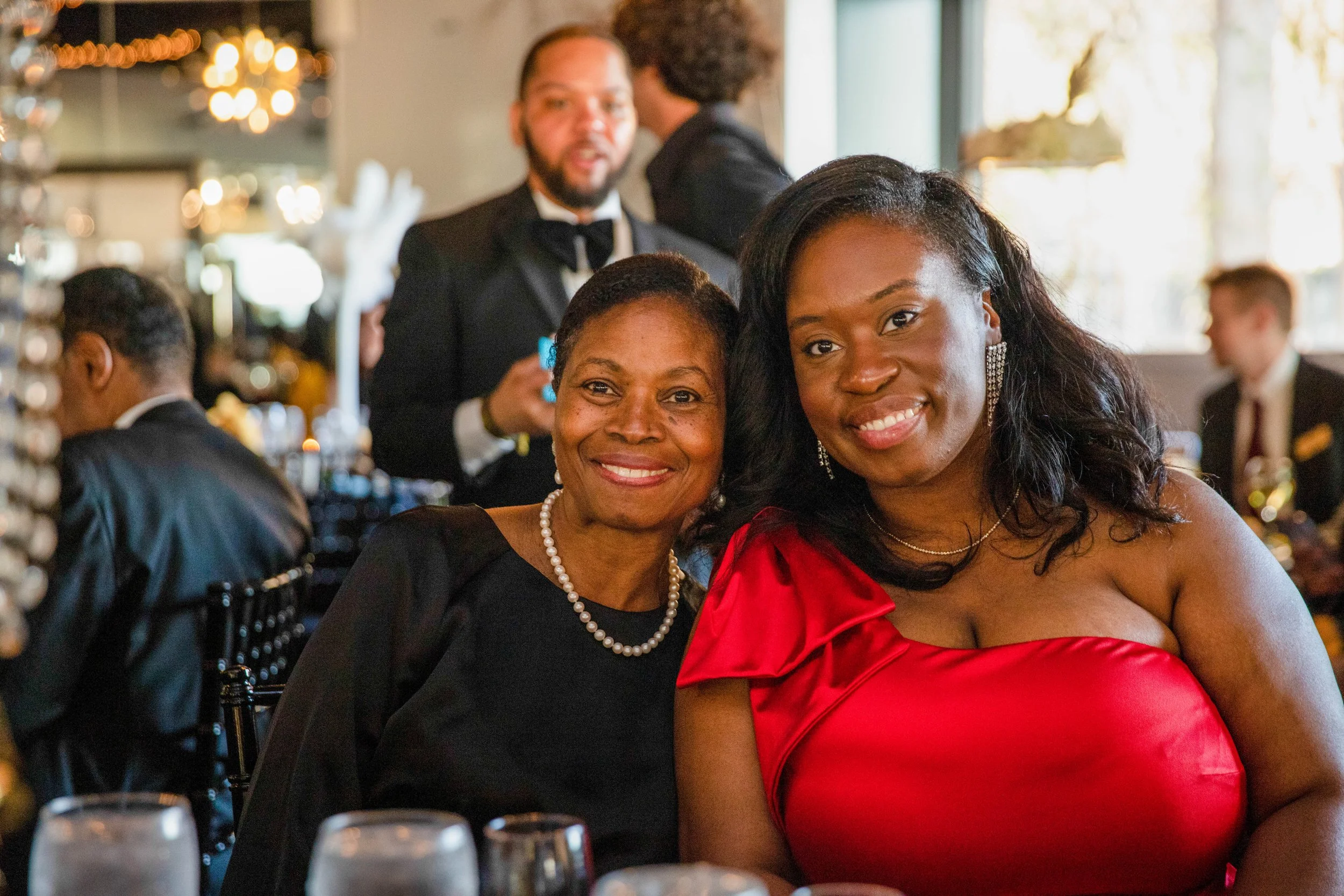 Two women, one older and one younger, smiling at a formal event, with the younger woman wearing a red dress and the older woman wearing a black dress and pearl necklace. In the background, a man in a tuxedo and other people are visible, with a blurre