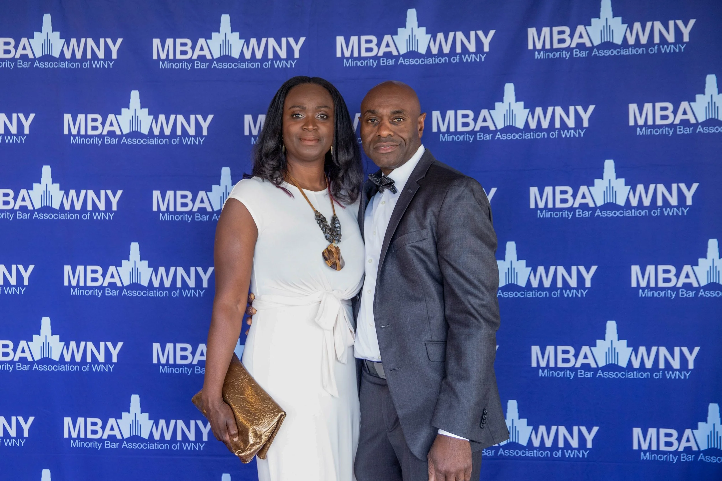 A man and woman standing together in formal attire in front of a blue backdrop with the logo for the Minority Bar Association of WNY. The woman is wearing a white dress and holding a gold clutch, while the man is wearing a dark suit with a bow tie.