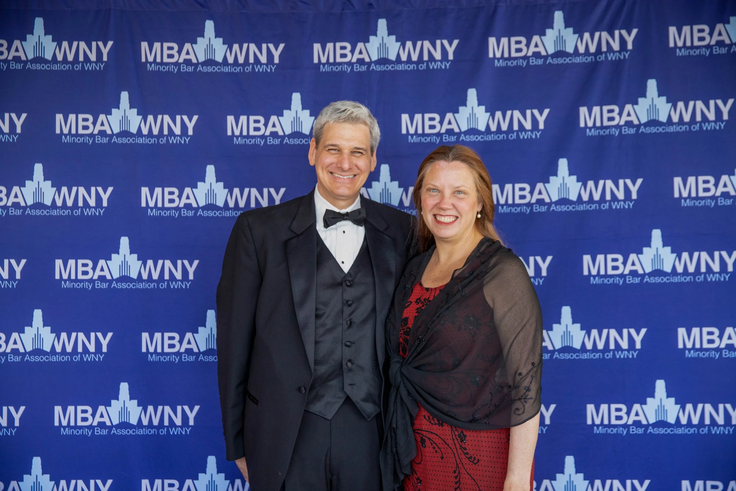 Two people, a man in a tuxedo and a woman in a red and black dress, smiling at a formal event with a blue backdrop that has the logo and text of the Minority Bar Association of WNY.