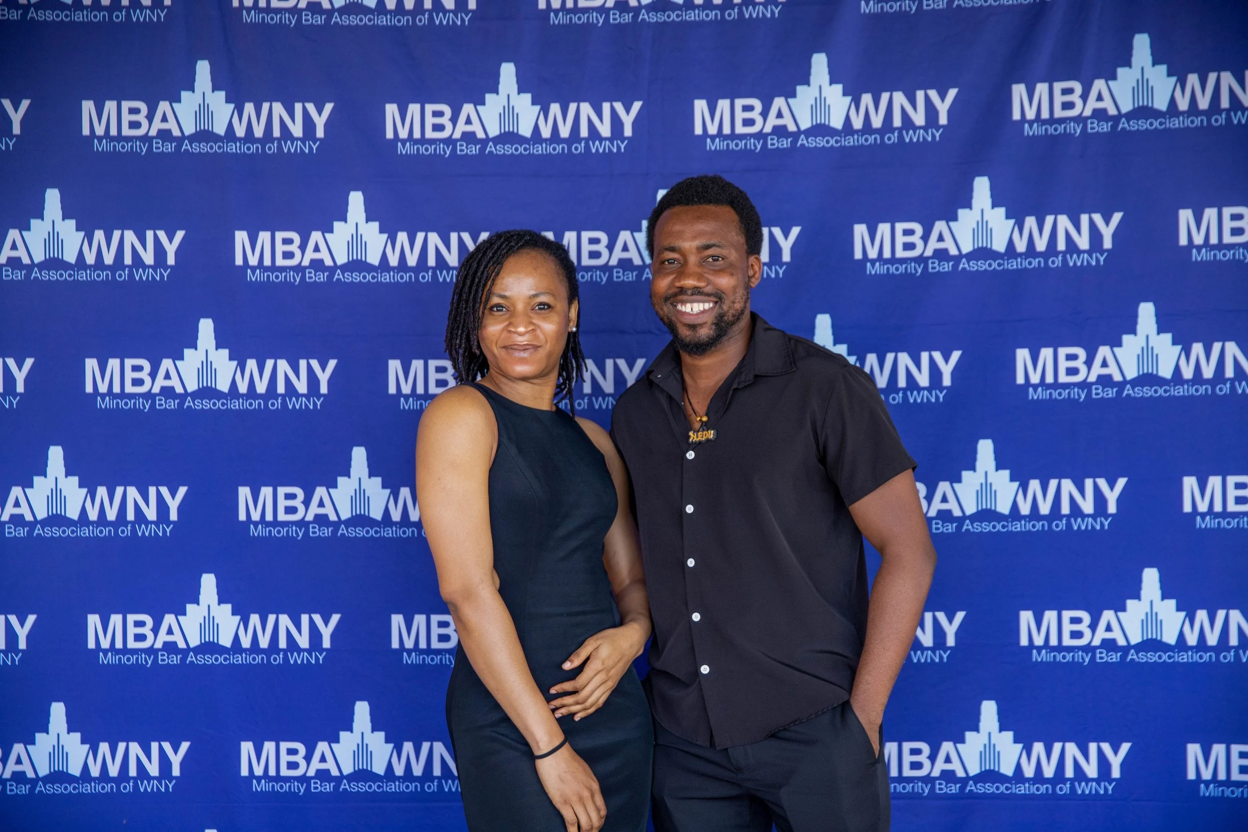 A man and woman standing together in front of a blue backdrop with the logo and text for the Minority Bar Association of WNY, smiling at the camera.