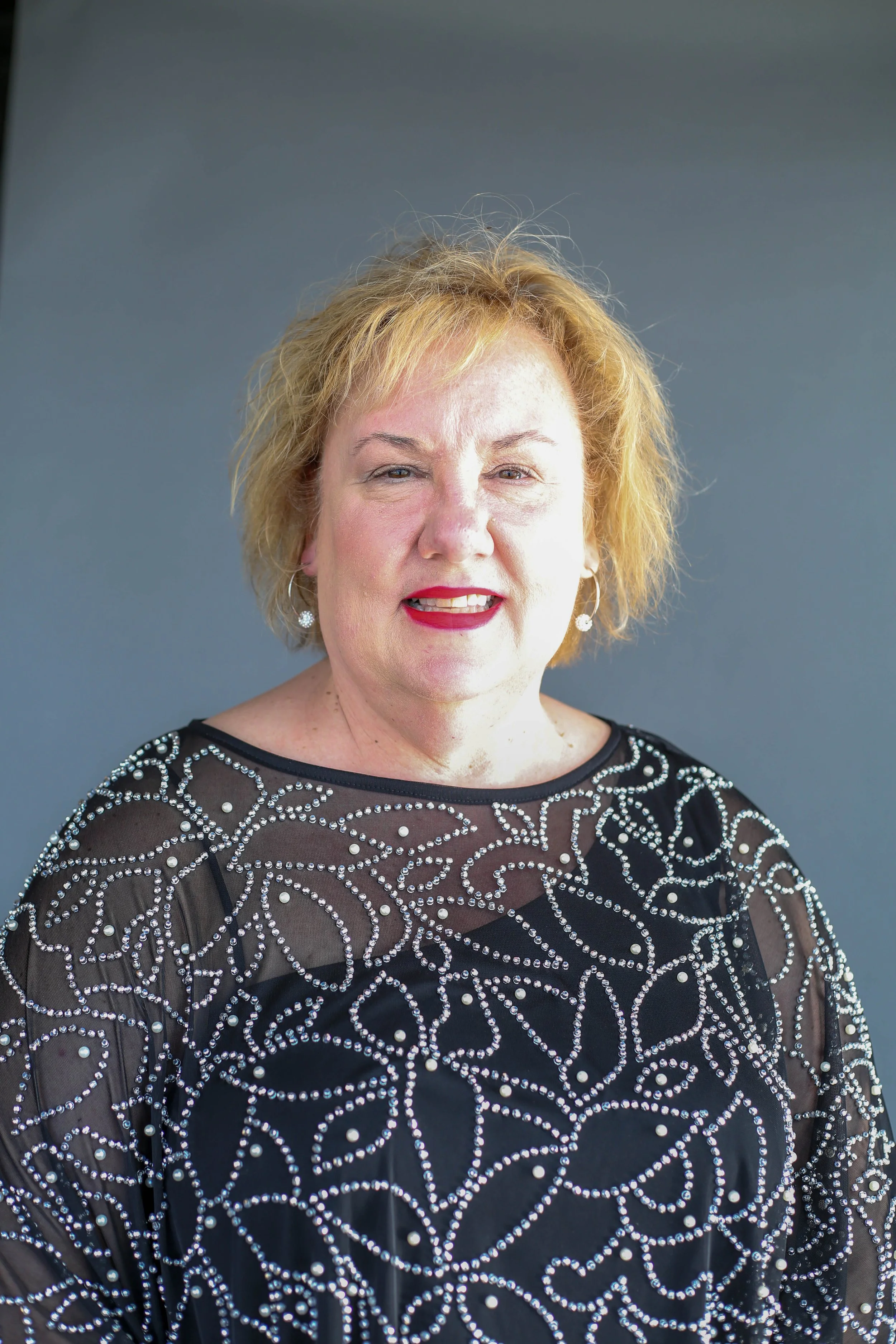 Close-up of a woman with light skin and curly blonde hair, smiling and wearing a black dress with intricate silver patterns and sheer fabric on the sleeves.