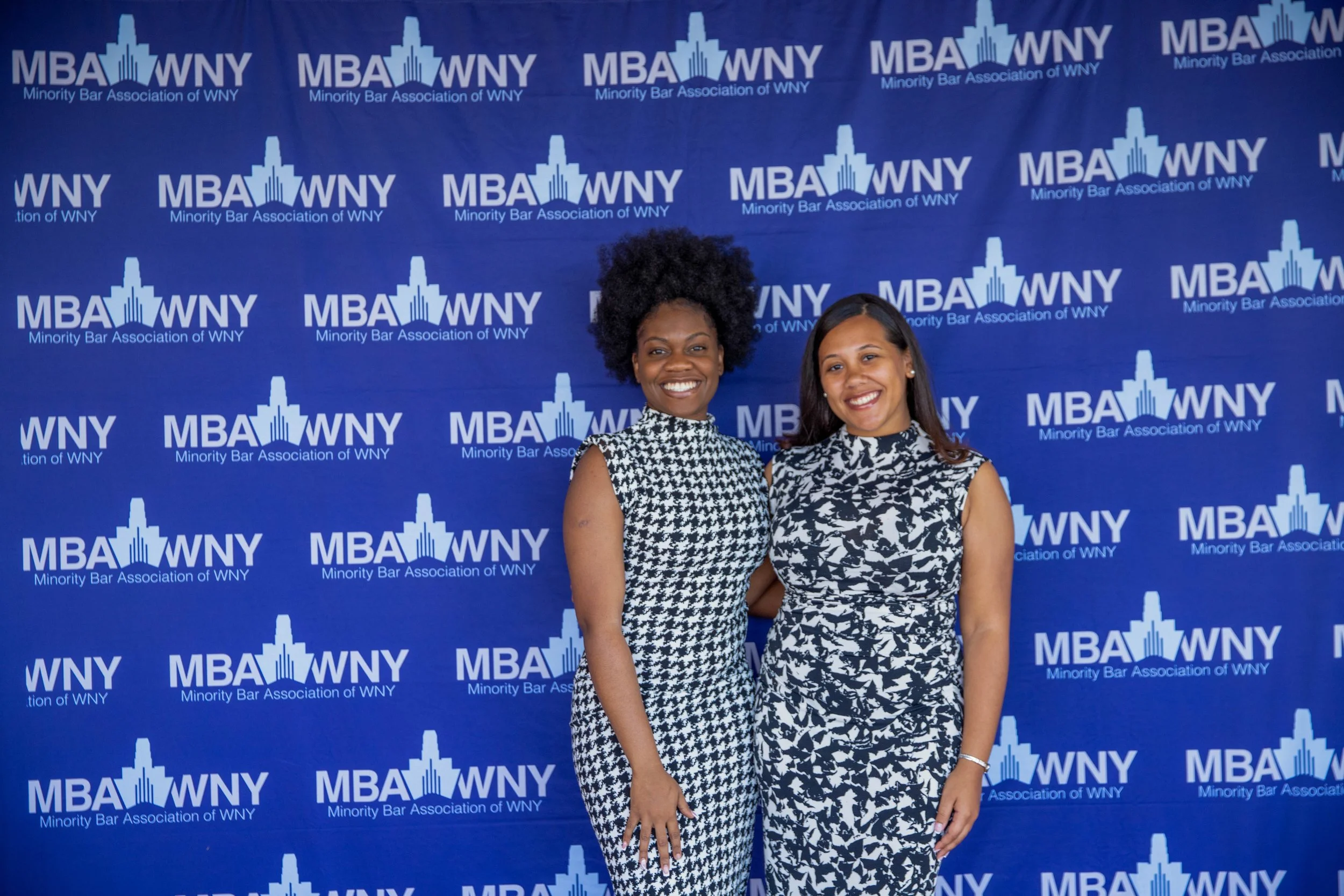 Two women standing in front of a blue backdrop with the logo and text for the Minority Bar Association of WNY, smiling at the camera. The woman on the left has dark skin and curly hair, wearing a black and white patterned dress. The woman on the righ