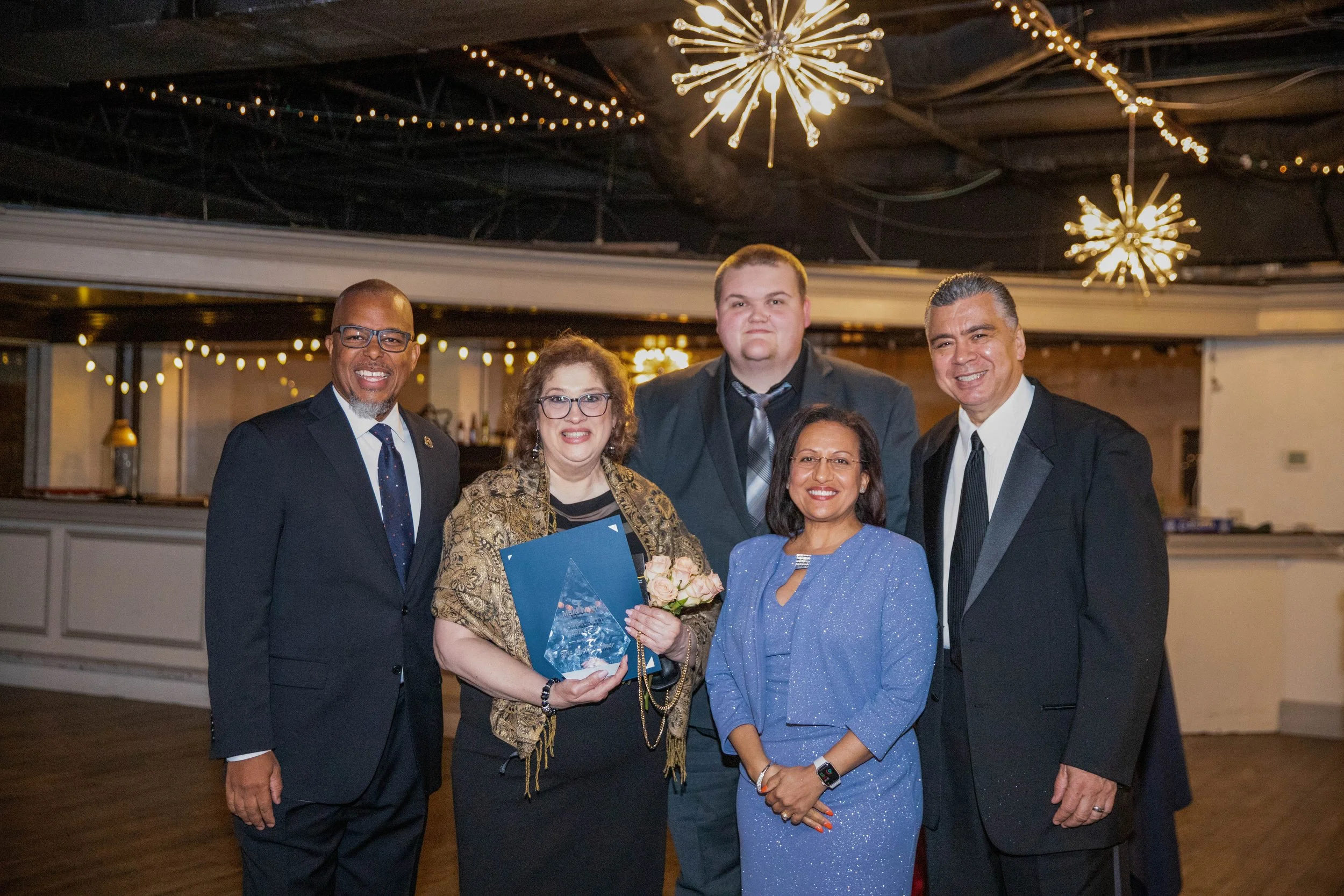 A group of five people dressed in formal attire standing together at an indoor event, with a woman in the center holding a bouquet of flowers and a glass award.