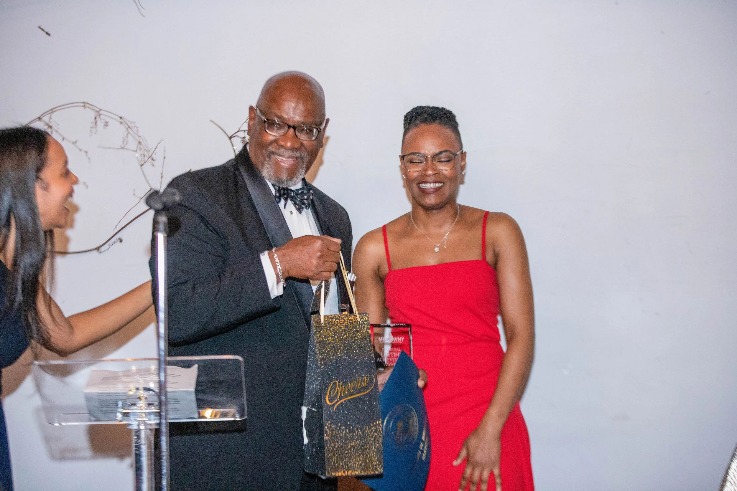 A group of people at an indoor event, with a man in a tuxedo and bowtie standing next to a woman in a red dress, both smiling and holding awards or gift bags. Others are partially visible around them.