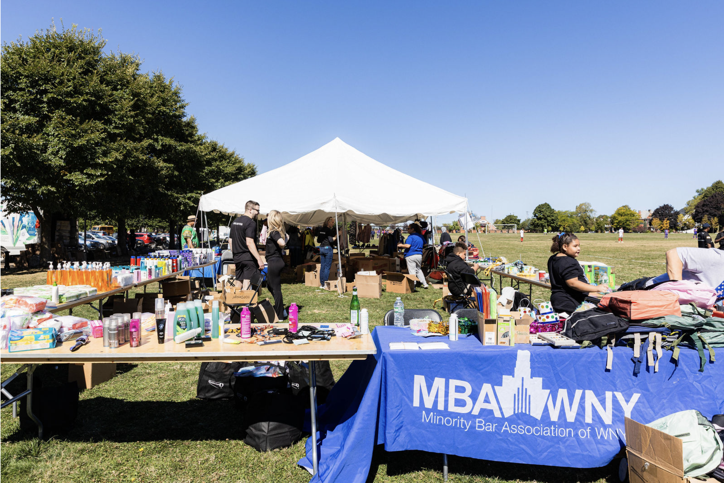 Outdoor community event with tables of various items like toiletries, clothing, and accessories under a white canopy tent. People are browsing and interacting, with a park and trees in the background on a sunny day.