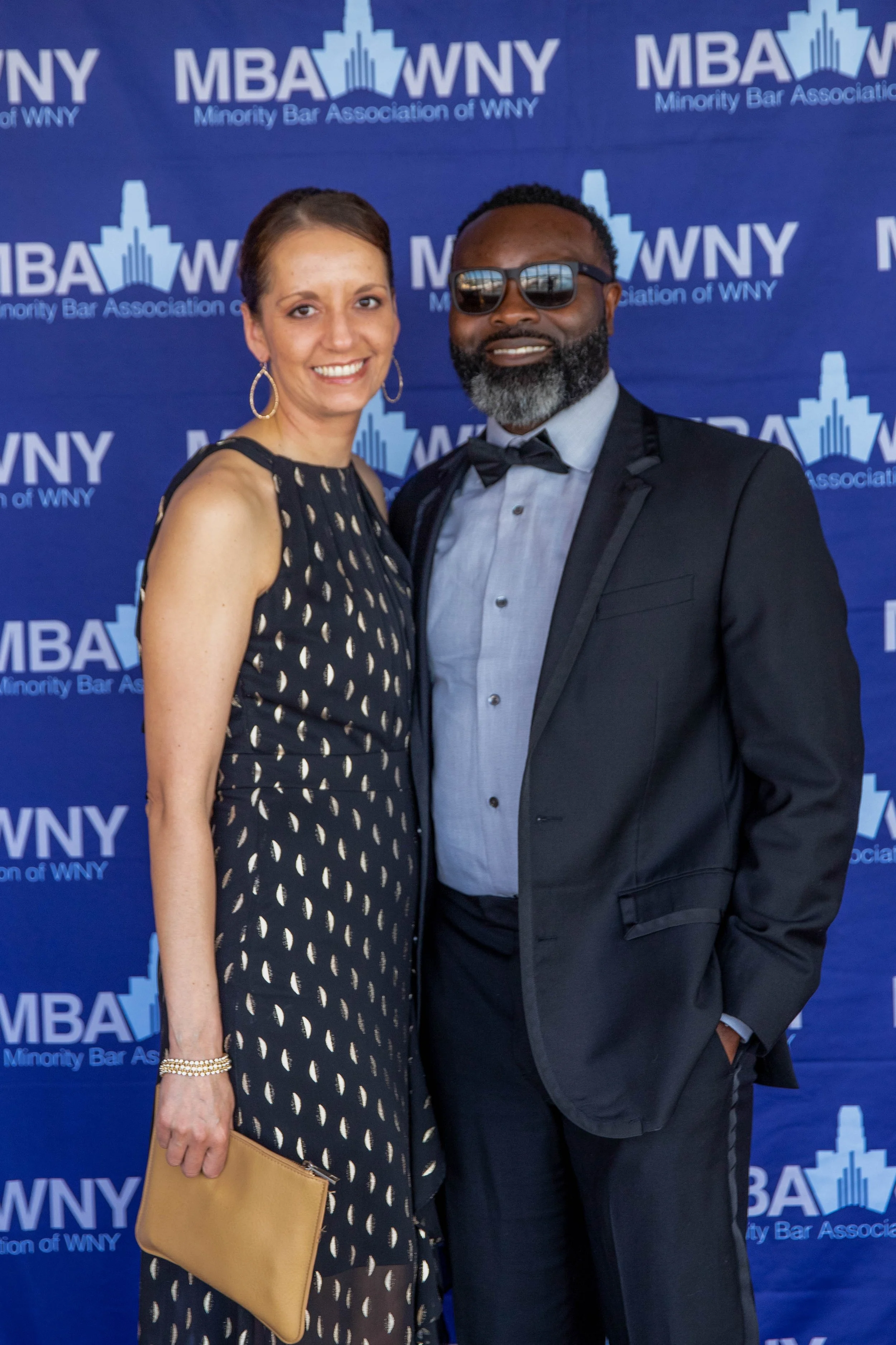 A man and woman dressed formally, smiling and standing together at an event, with a blue backdrop featuring the logo of MBA WNY, Minorities Bar Association of Western New York.