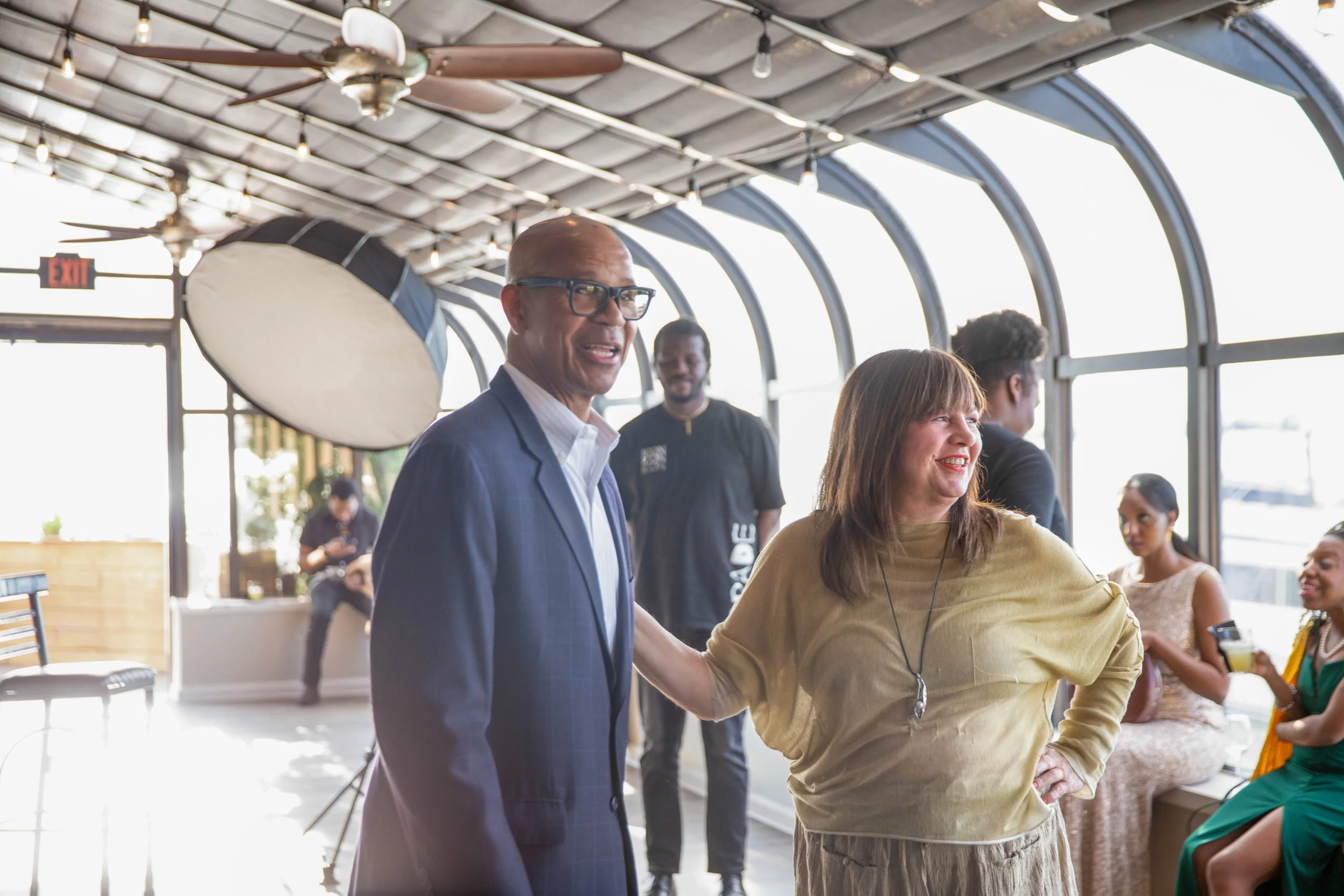 A group of diverse people, including a man in a suit and glasses and a woman in a yellow top, are smiling and engaging with others at a social event indoors with large arched windows and a ceiling fan.