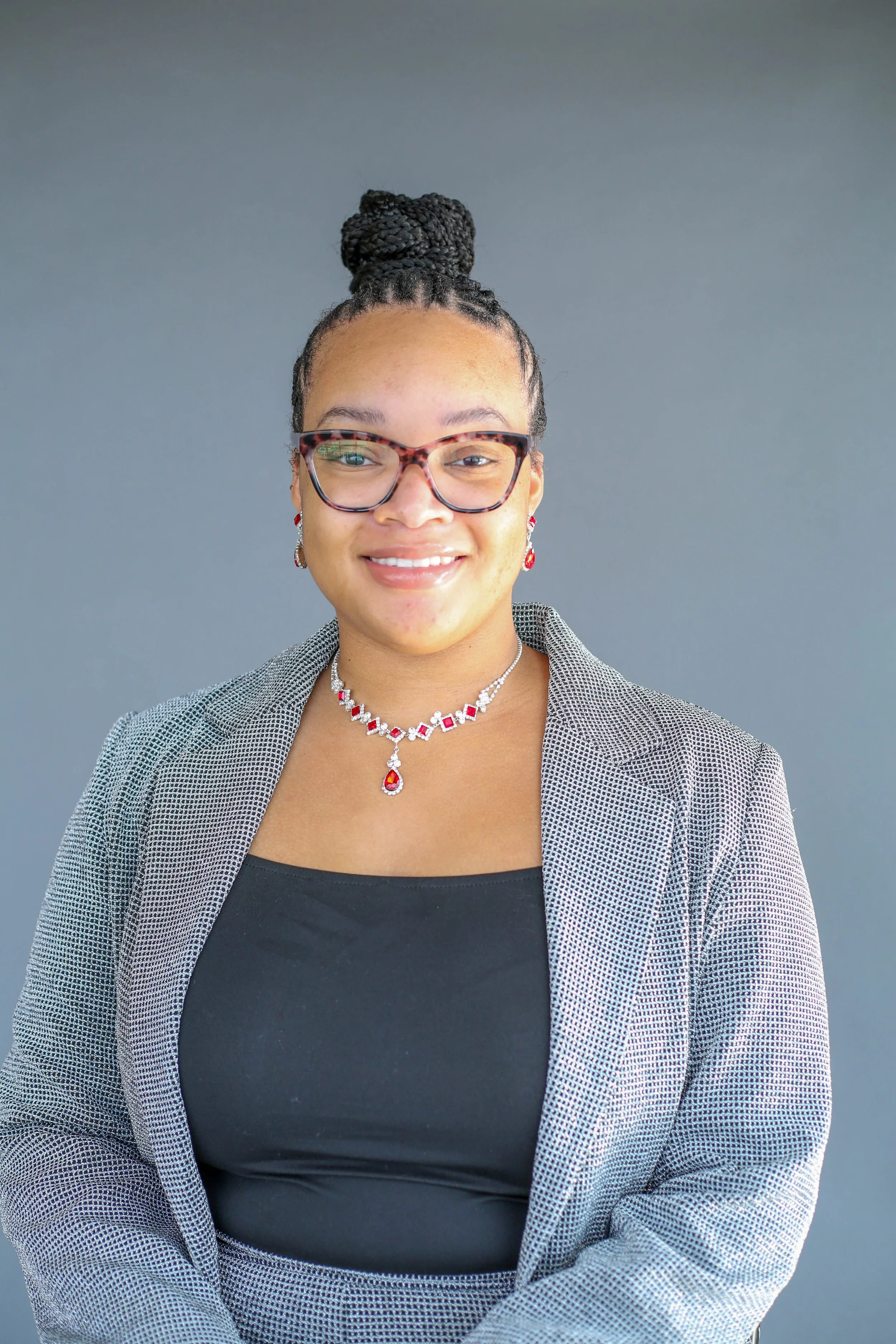 A woman with glasses and earrings wearing a plaid blazer over a black top, smiling against a plain gray background.
