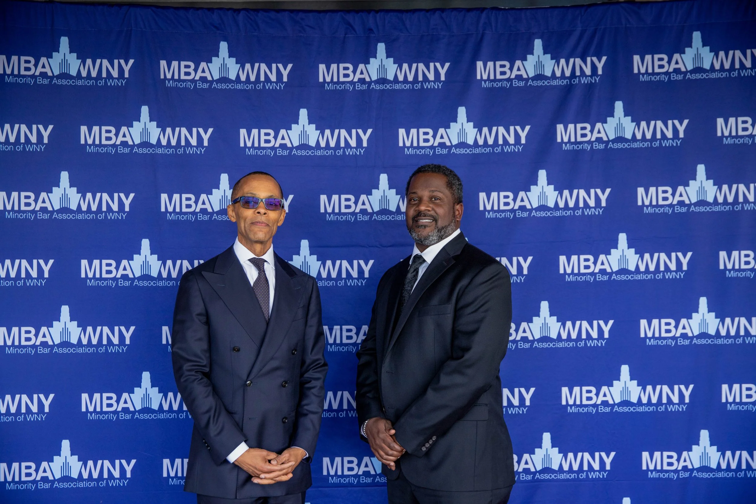 Two men in suits standing in front of a blue backdrop with white lettering that reads "MBA WNY Minor League Bar Association of WNY" and features a skyline logo. One man wears glasses and a tie, the other has a beard and no glasses.