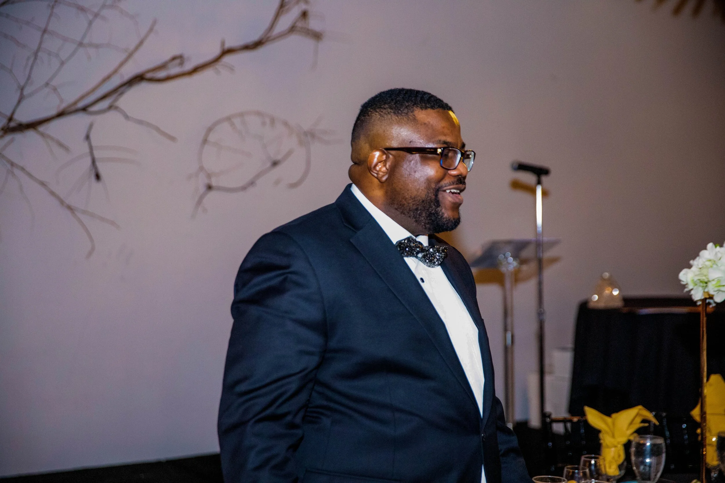 A man in a tuxedo with glasses and a bow tie smiling at a formal event. There are table decorations including a flower arrangement and yellow napkins in the background.